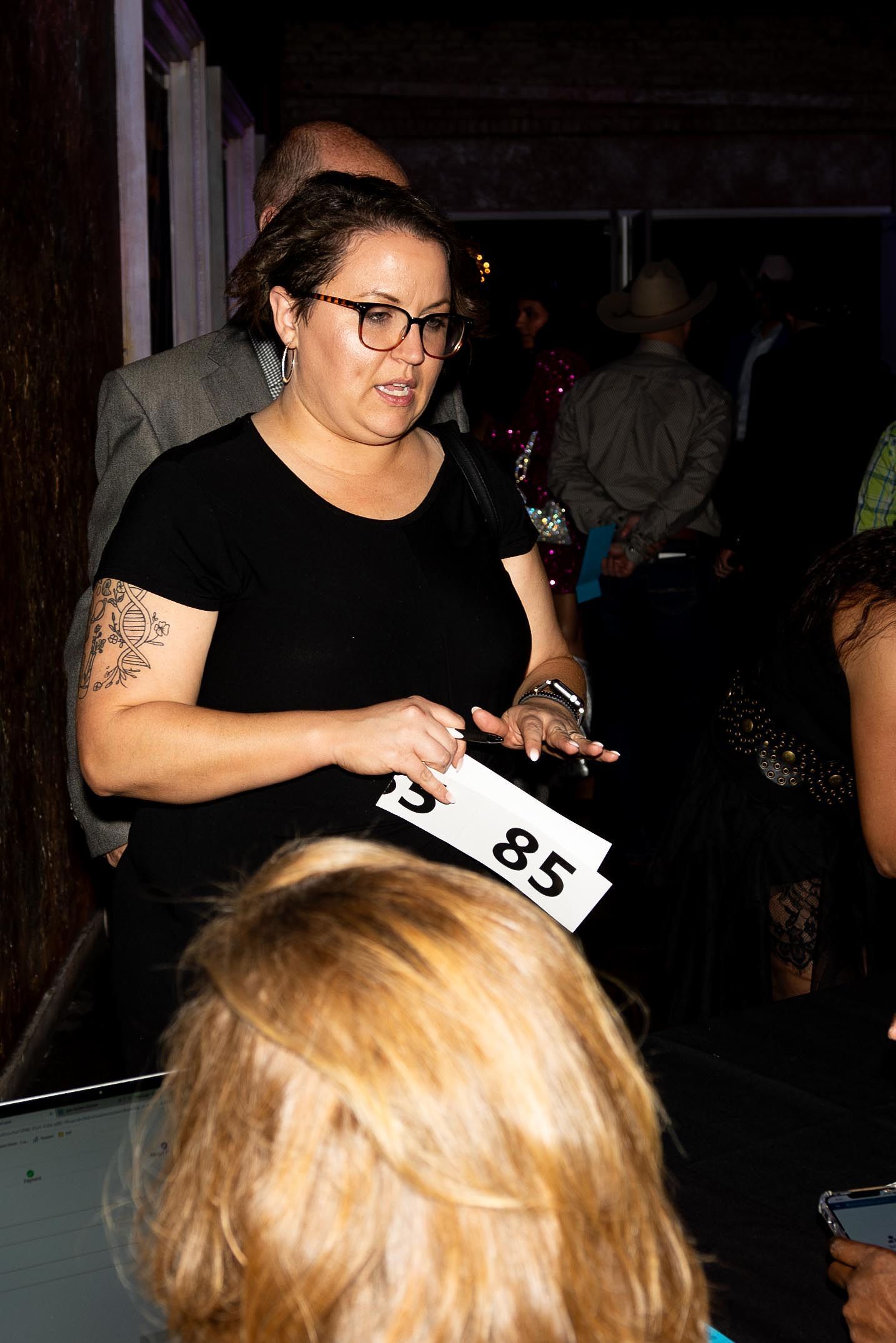Woman with glasses holding a bidding paddle, in an event setting.