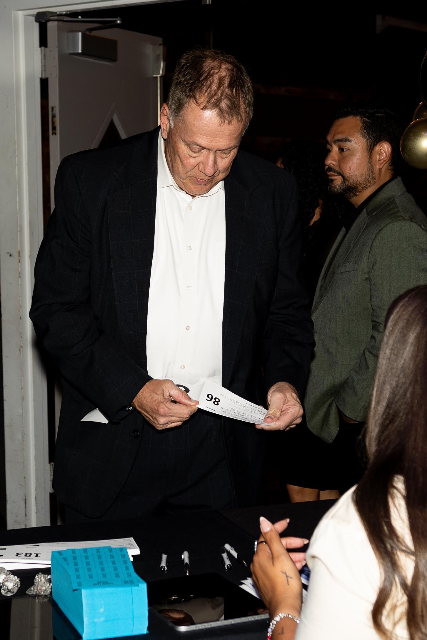 Man in a suit examining paper, standing near a table, with another person in the background.