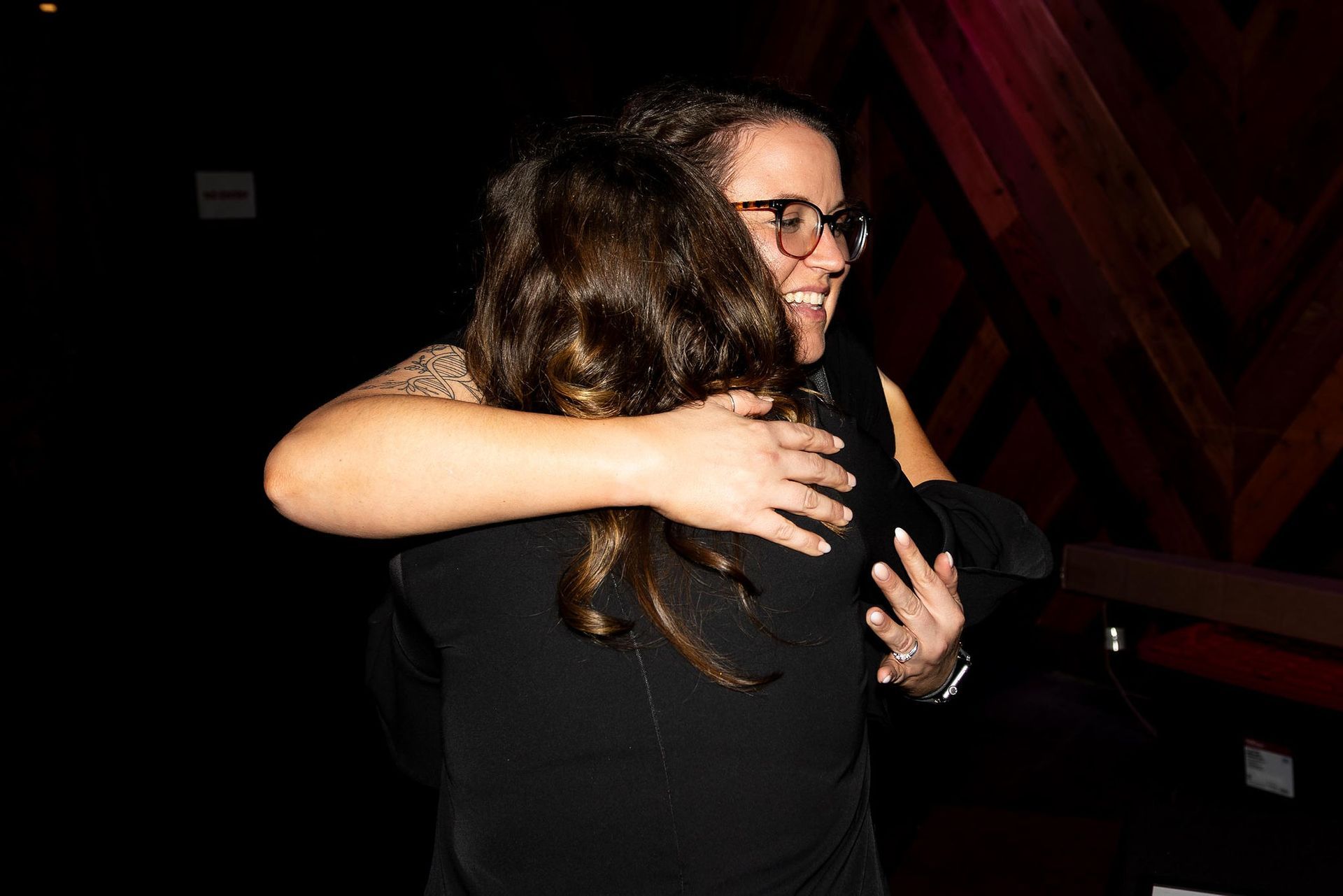 Two women embracing indoors. One wears glasses, smiles, other's back to camera. Dark setting, wooden background.