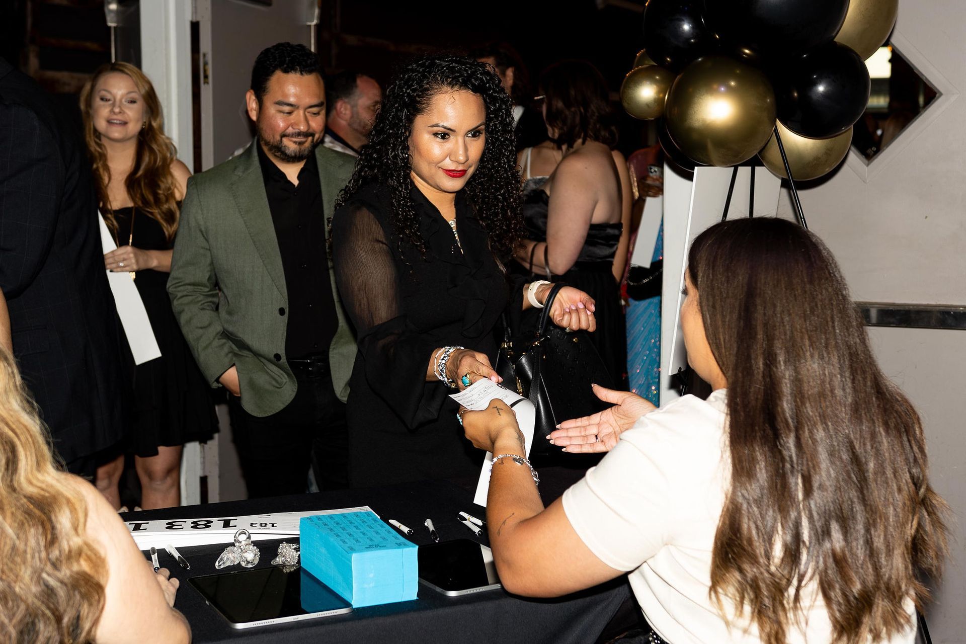 Woman in black dress pays at a table with jewelry, others look on, balloons overhead.