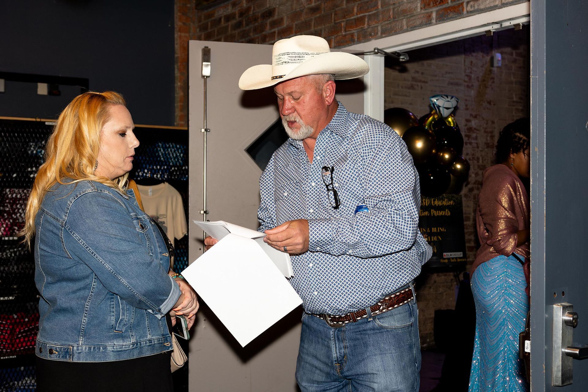 Woman in jean jacket listens to man in cowboy hat reading papers at a doorway.