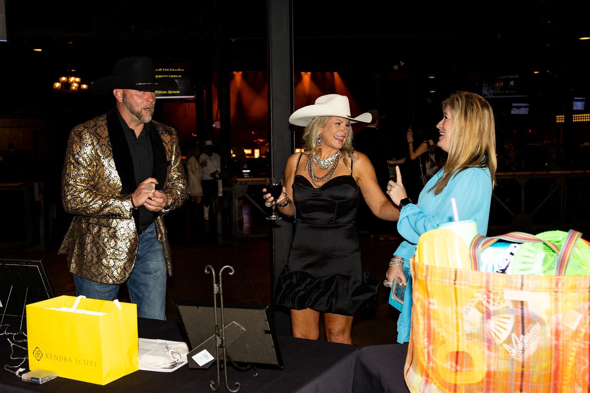 People at an event. Man in gold jacket, woman in black dress, woman in blue top, table with items.