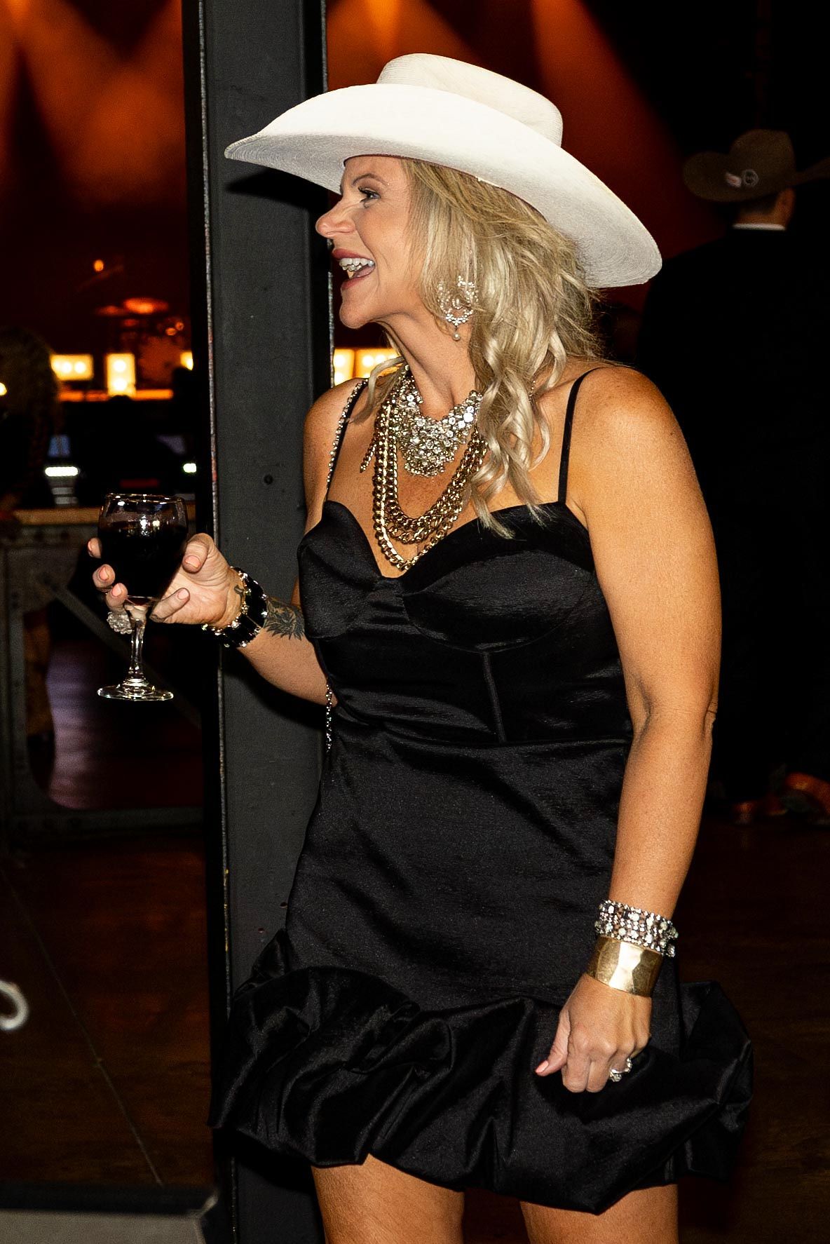 Woman in a black dress and white hat holds wine glass, smiling. Indoors, appears to be at an event.