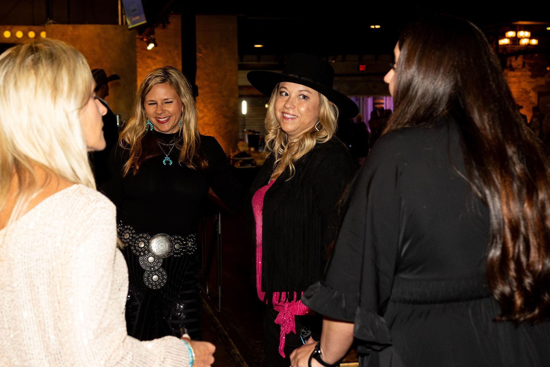 Four women talking at a dimly lit event. One wears a black hat and pink fringe. Others have blonde and brown hair.