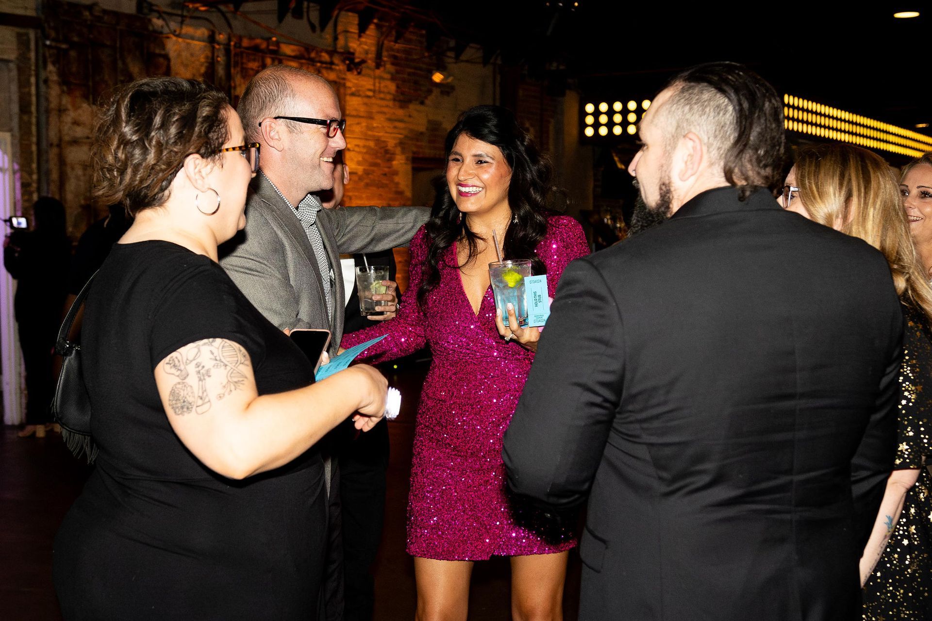 People in a party setting, smiling and talking, some holding drinks. Well-lit room, dark clothing, pink dress.
