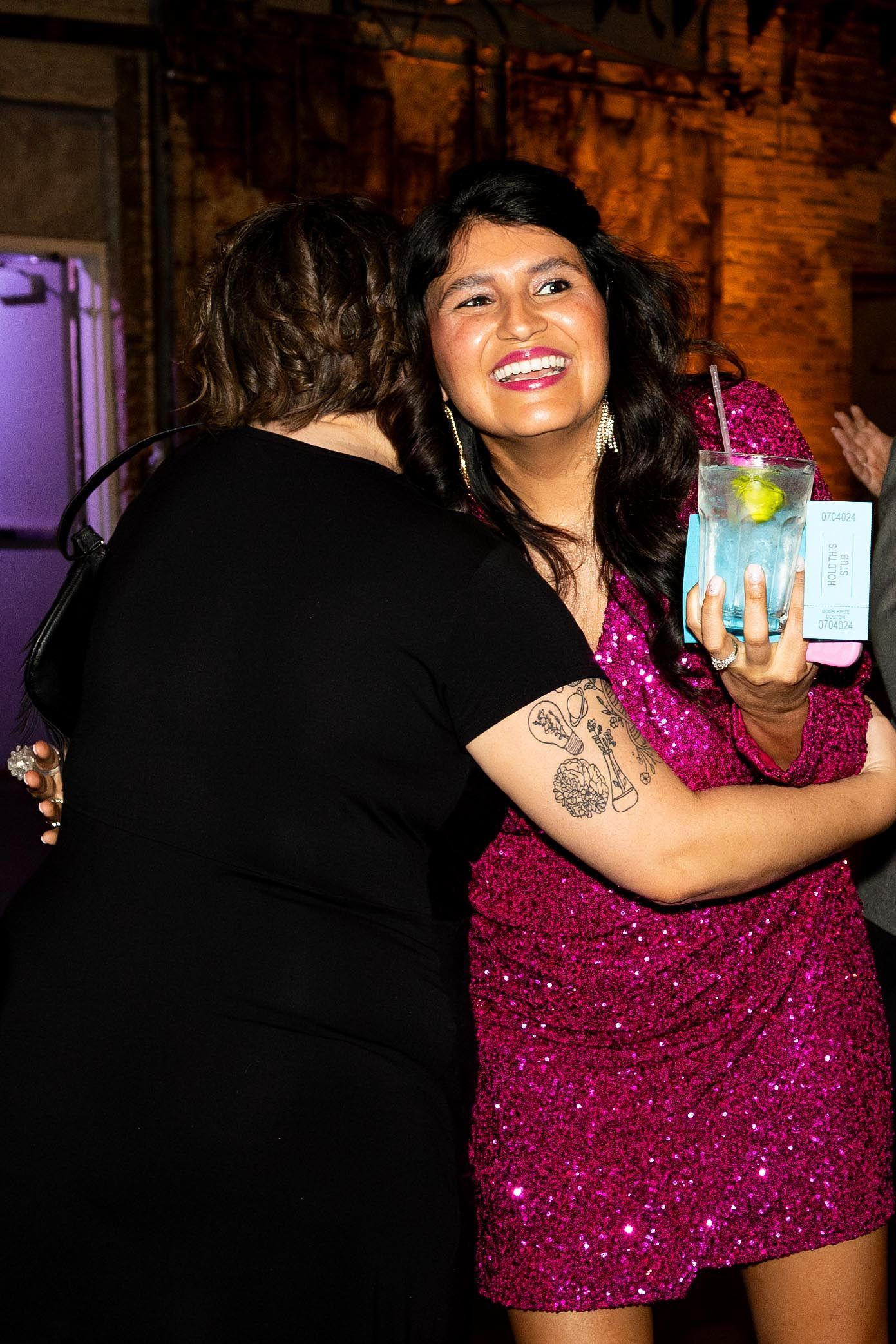 Woman in pink sequin dress hugging another, holding a drink, smiling.