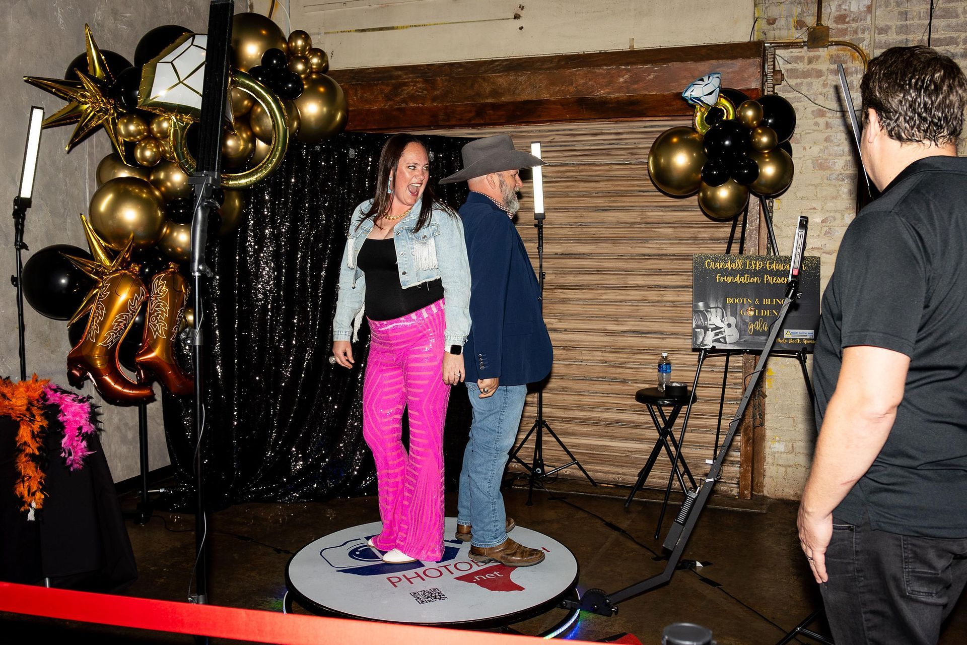 Woman in pink pants and man in a hat pose back-to-back on a rotating platform; a man films them.