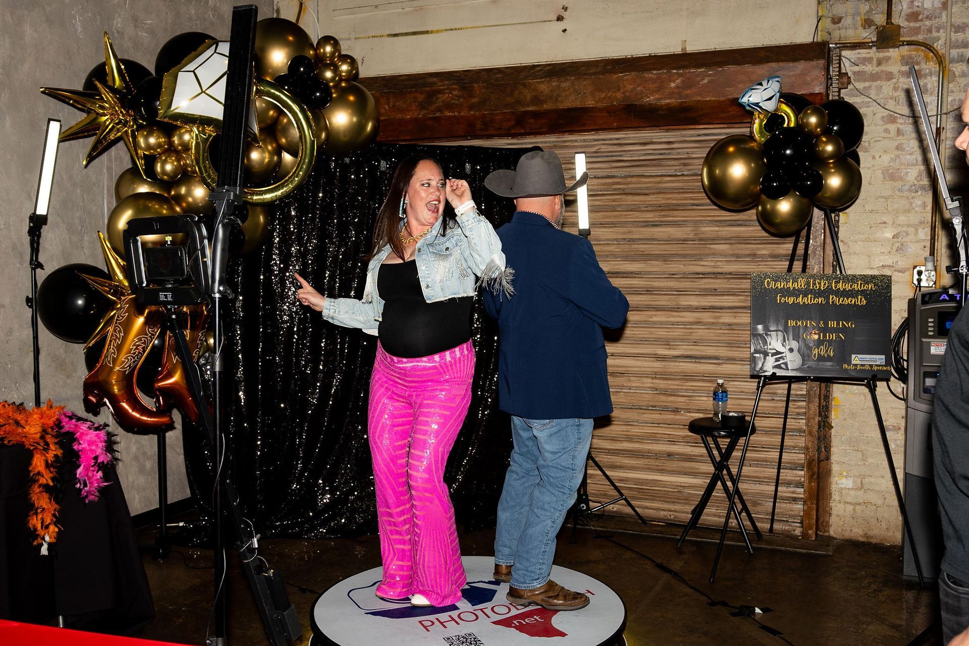 Woman in pink pants and man in cowboy hat pose on a photo booth platform, black and gold balloons in background.