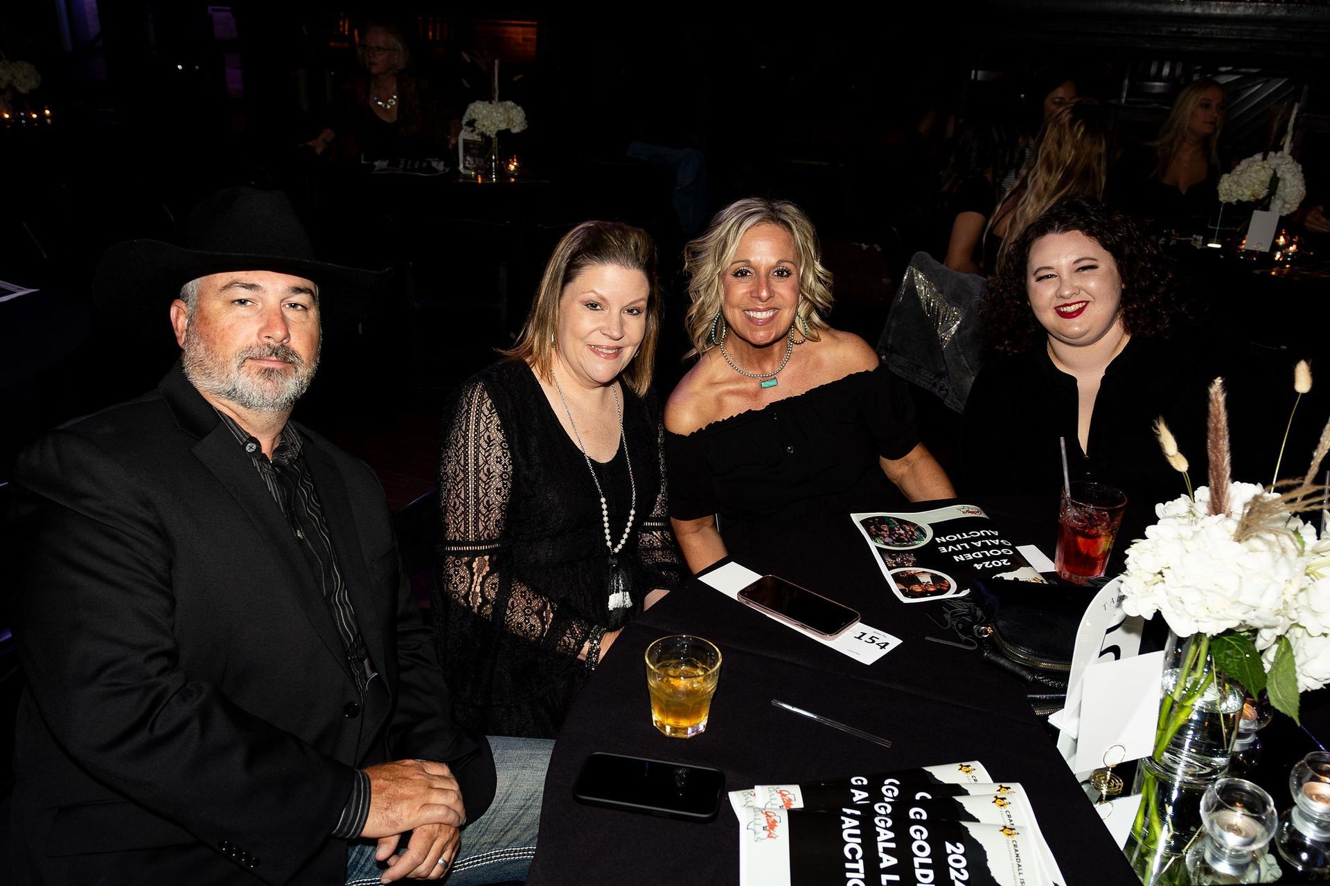 Four people at a dark event, seated at a table with drinks and a floral centerpiece.