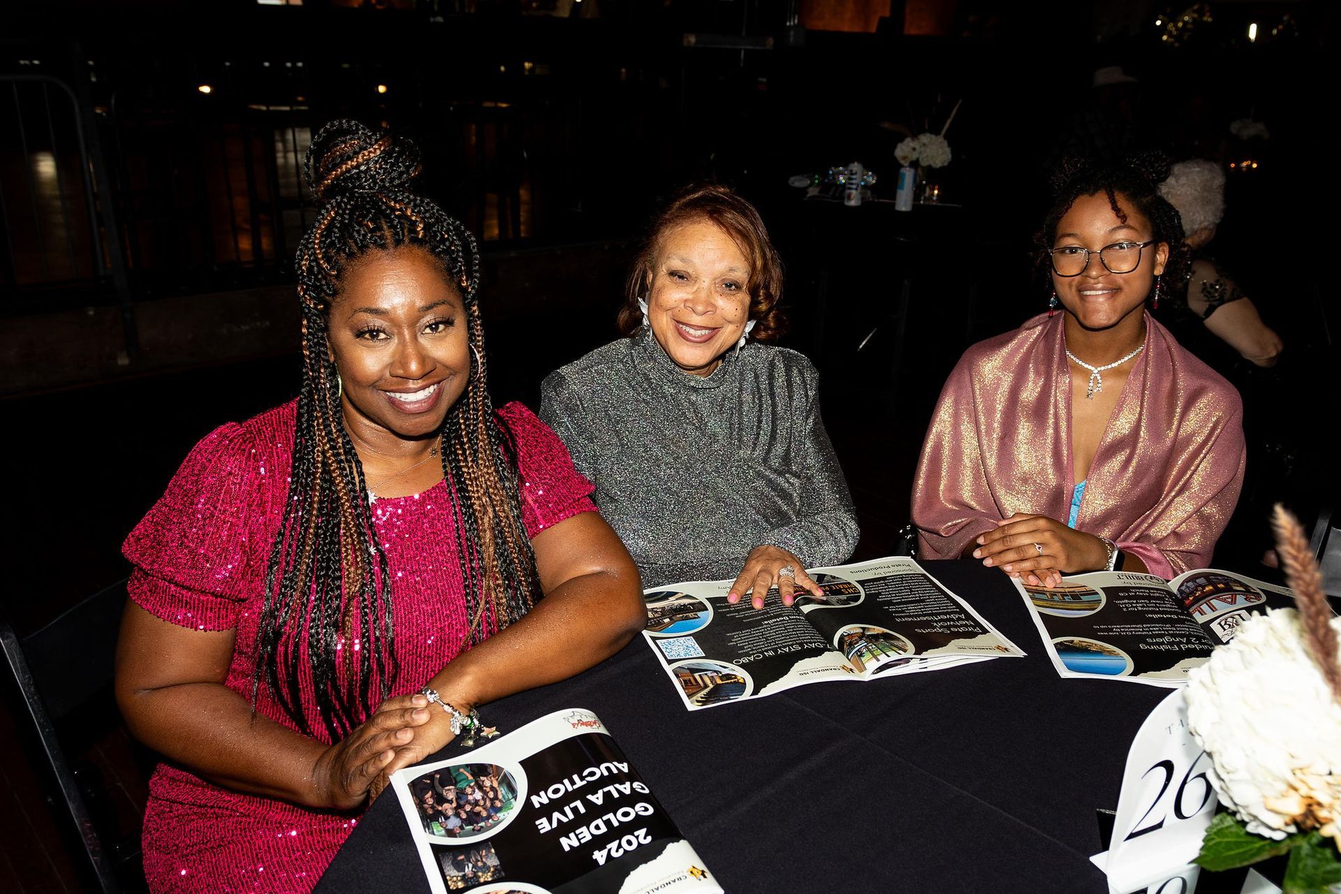 Three women smiling at a table with brochures. One wears red, one silver, one pink, in a dark room.