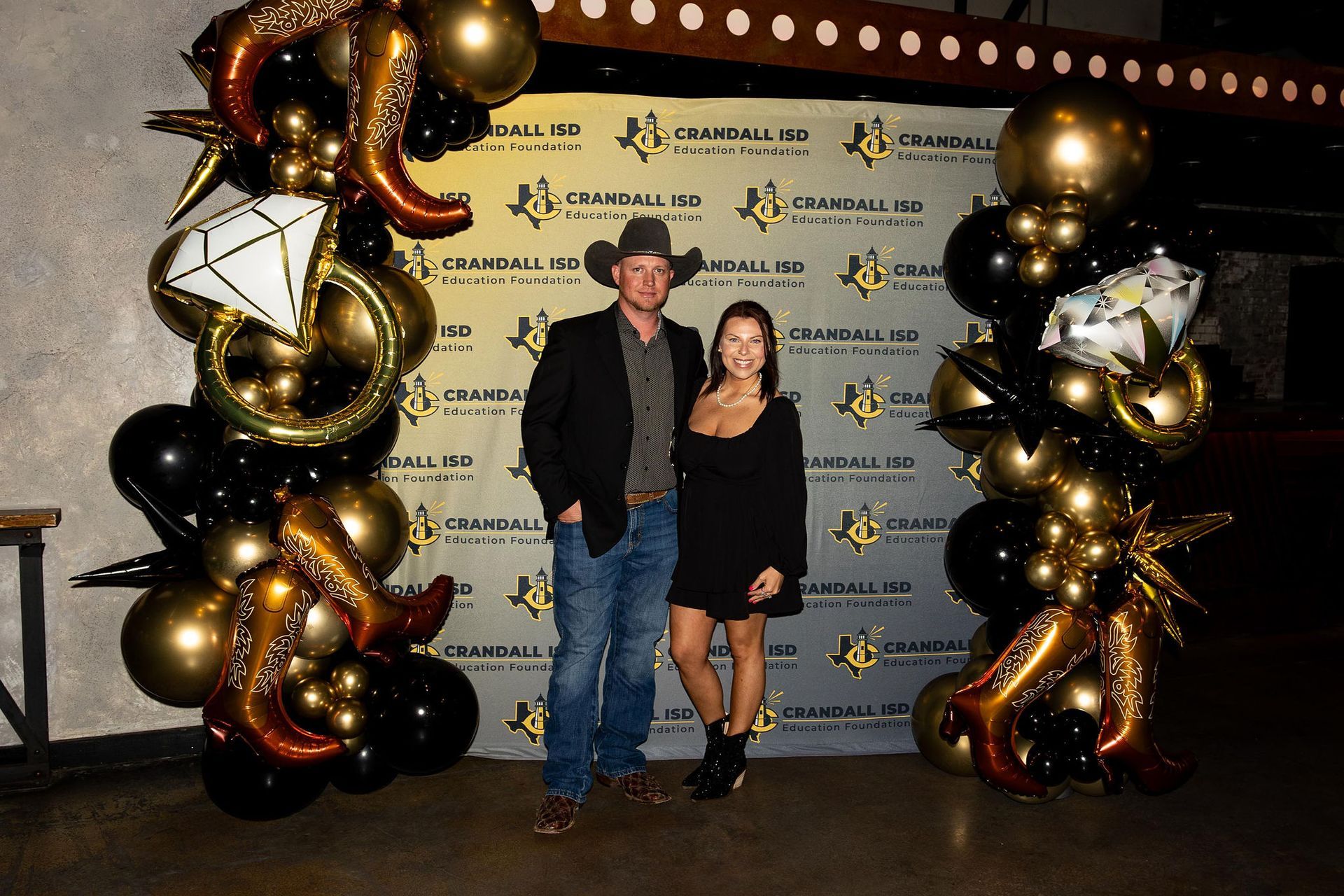 A couple poses in front of a backdrop with balloons; man in cowboy hat, woman in black dress.