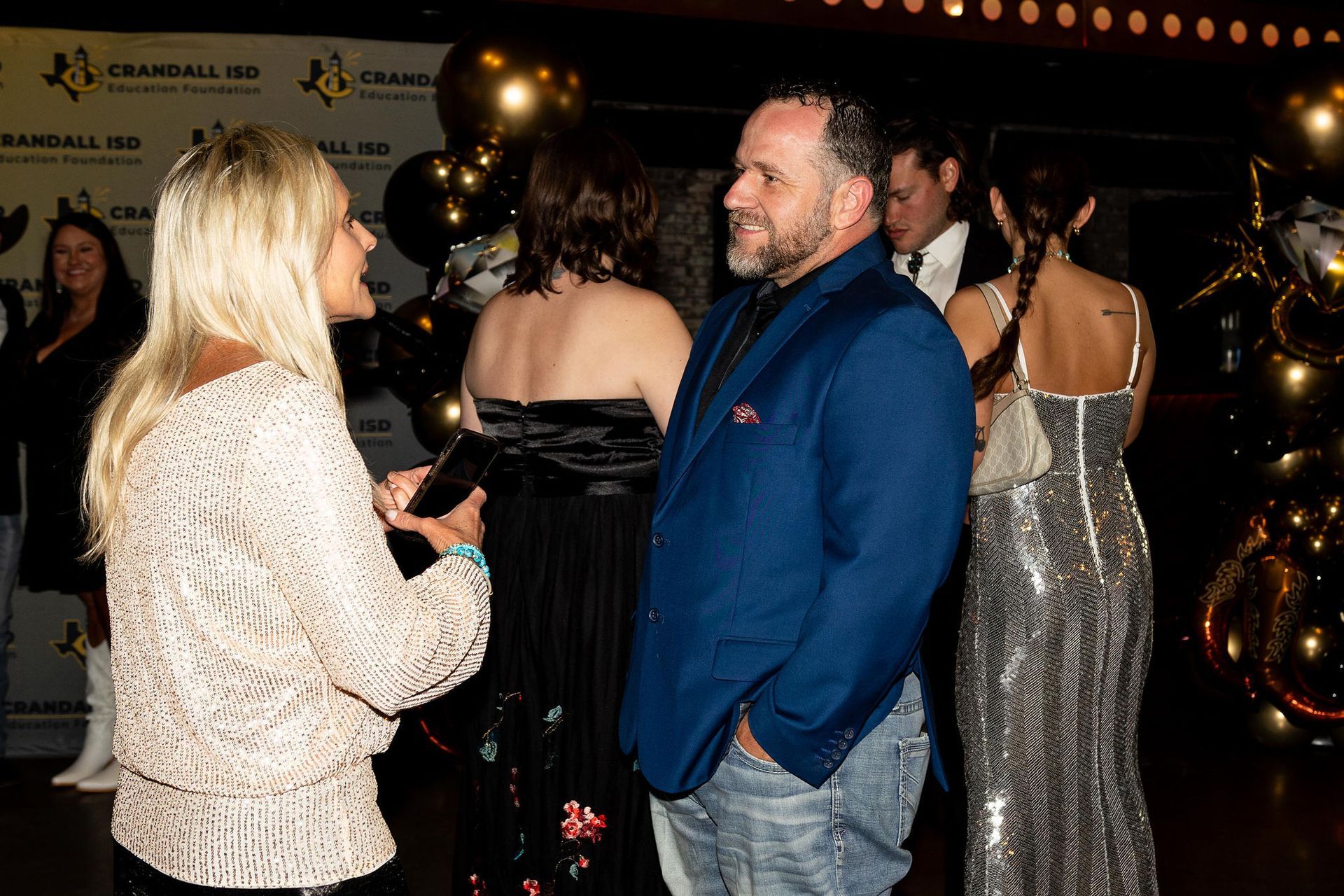People in formal attire at an event. Woman in lace top talks to a man in a blue blazer. Balloons in background.