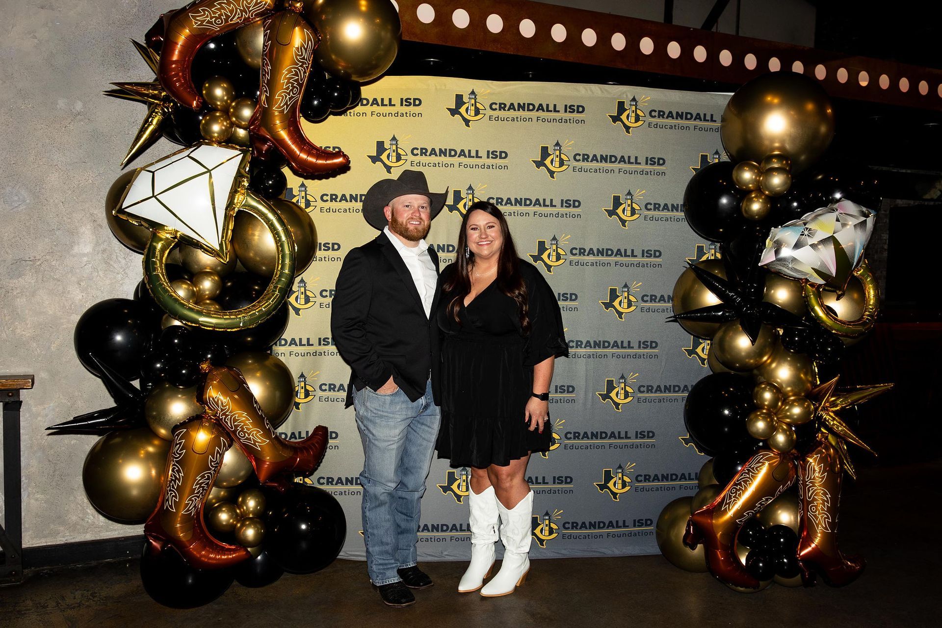 Couple posing in front of a branded backdrop, framed by black, gold, and cowboy boot balloons.