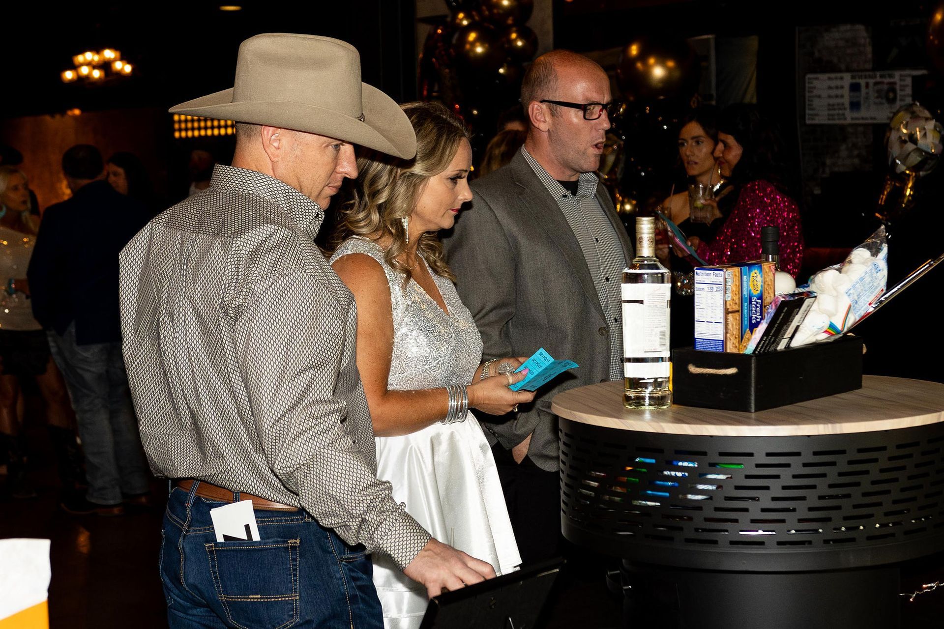 People at an event near a bar with a bottle of alcohol, a woman in a sparkly dress, and a man in a cowboy hat.