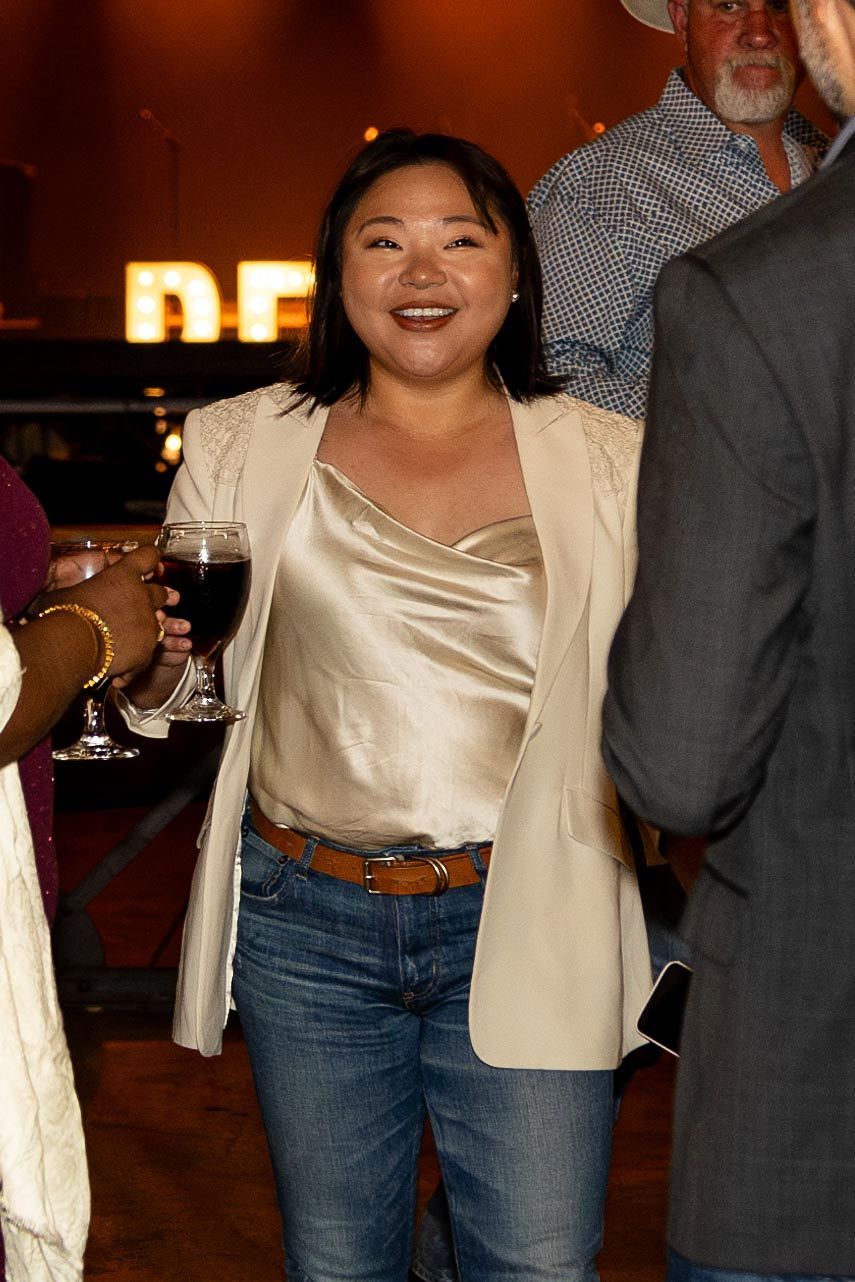 Woman with a glass of wine, smiling. Wearing a cream blazer, satin top, and jeans at an event.