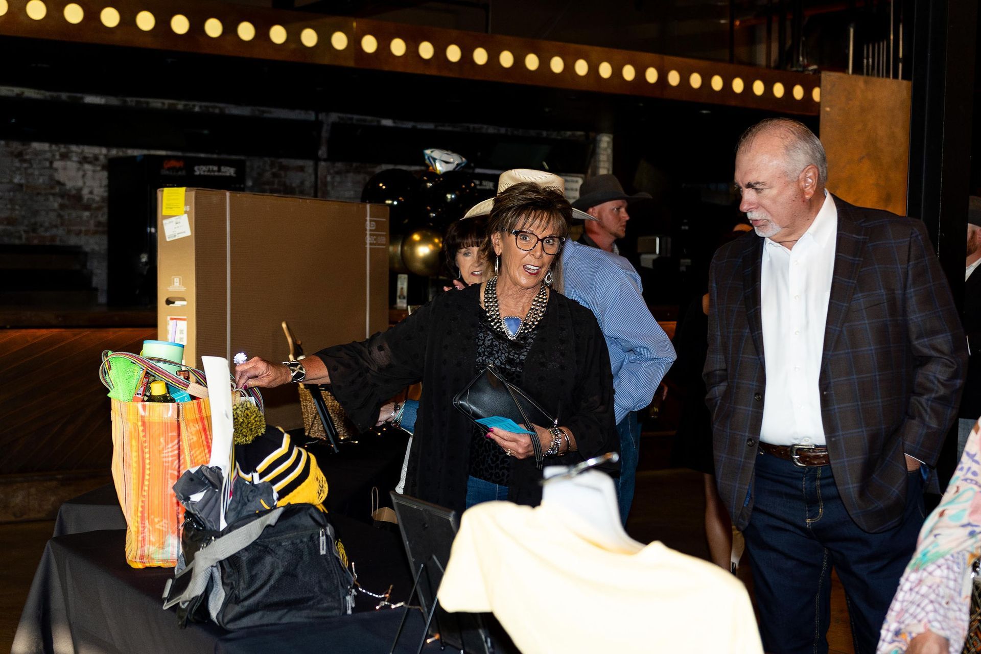 A woman pointing at auction items on a table, man beside her, in a dimly lit event space.