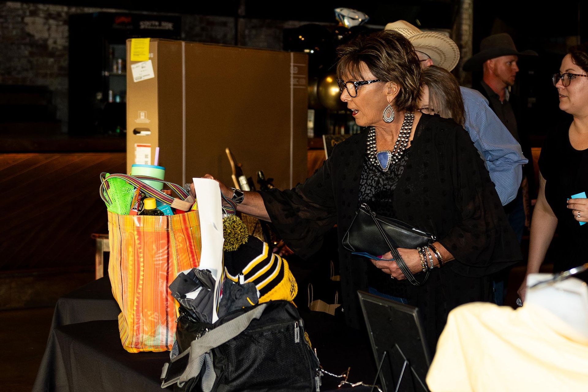 Woman at table with items, reaching. Others nearby, brick background, event at night.