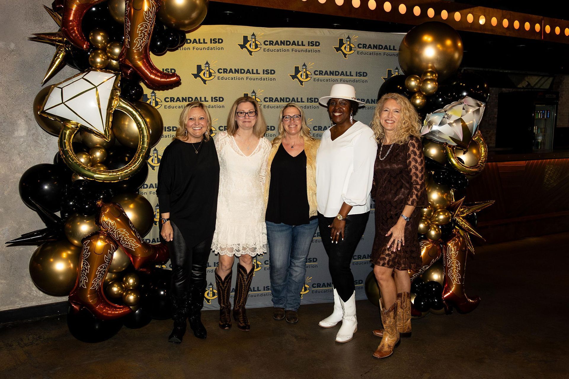 Five women in a celebratory setting with balloons, posing. One wears a white cowboy hat and boots. Gold, black, and brown.