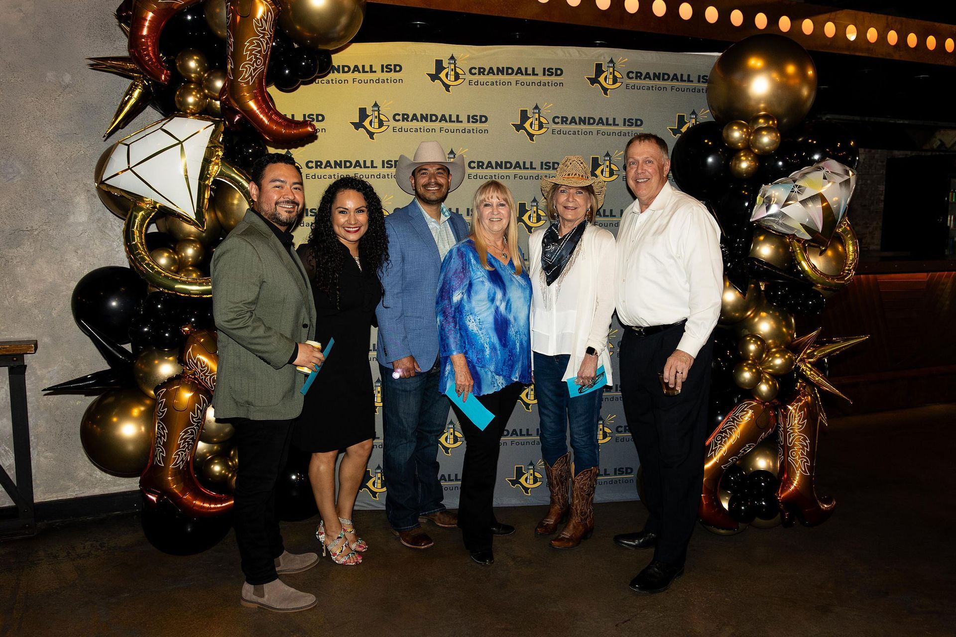 Group of people at an event posing in front of a backdrop with gold and black balloons.