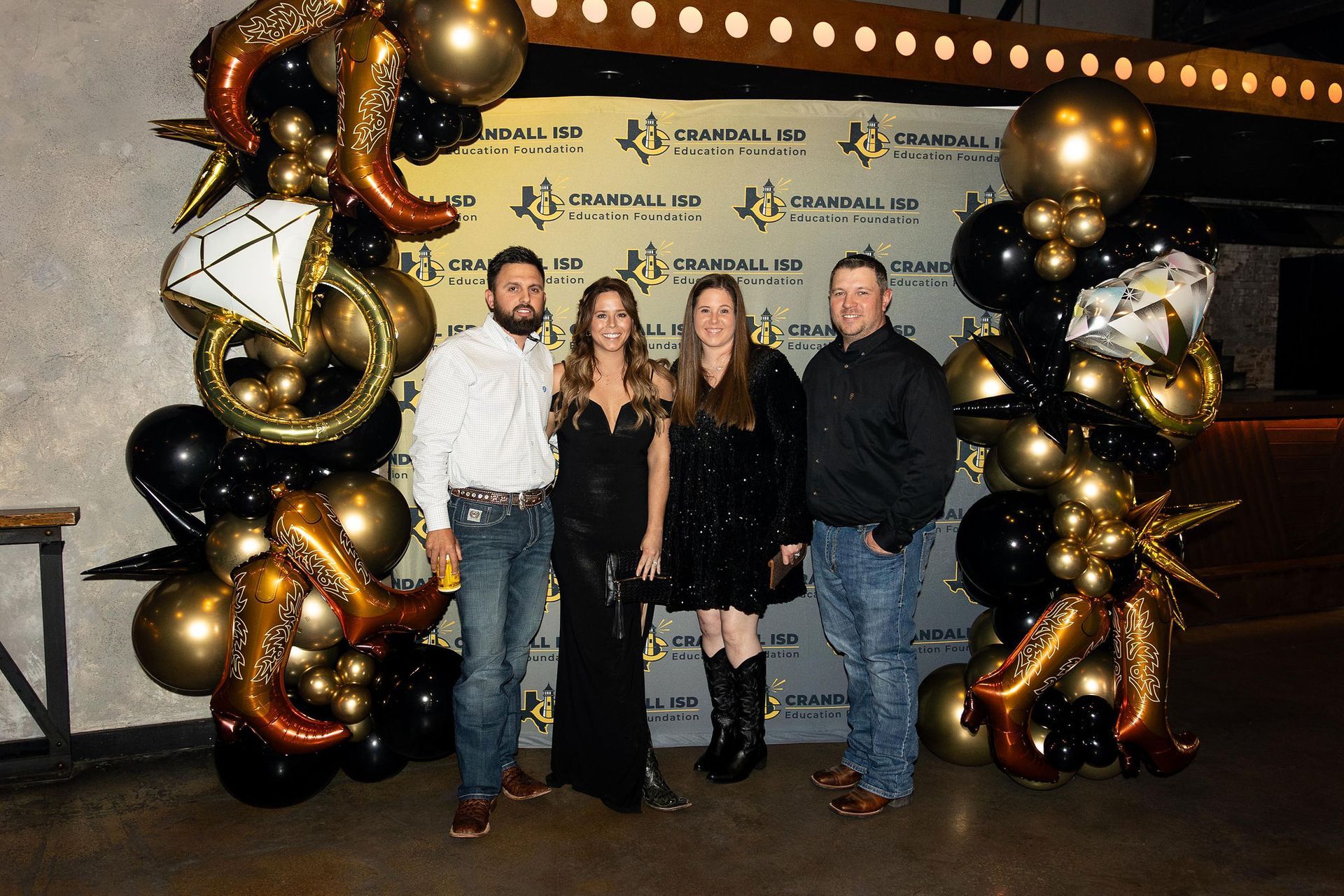 Four people pose in front of a backdrop with black and gold balloons at a party.