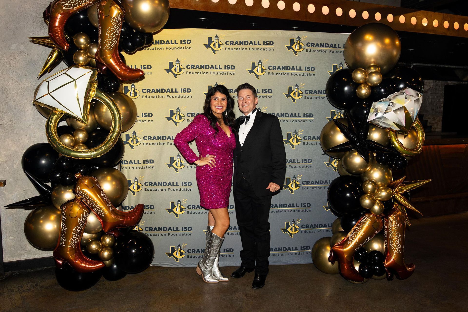 Couple poses for photo in front of a branded backdrop with gold and black balloons.