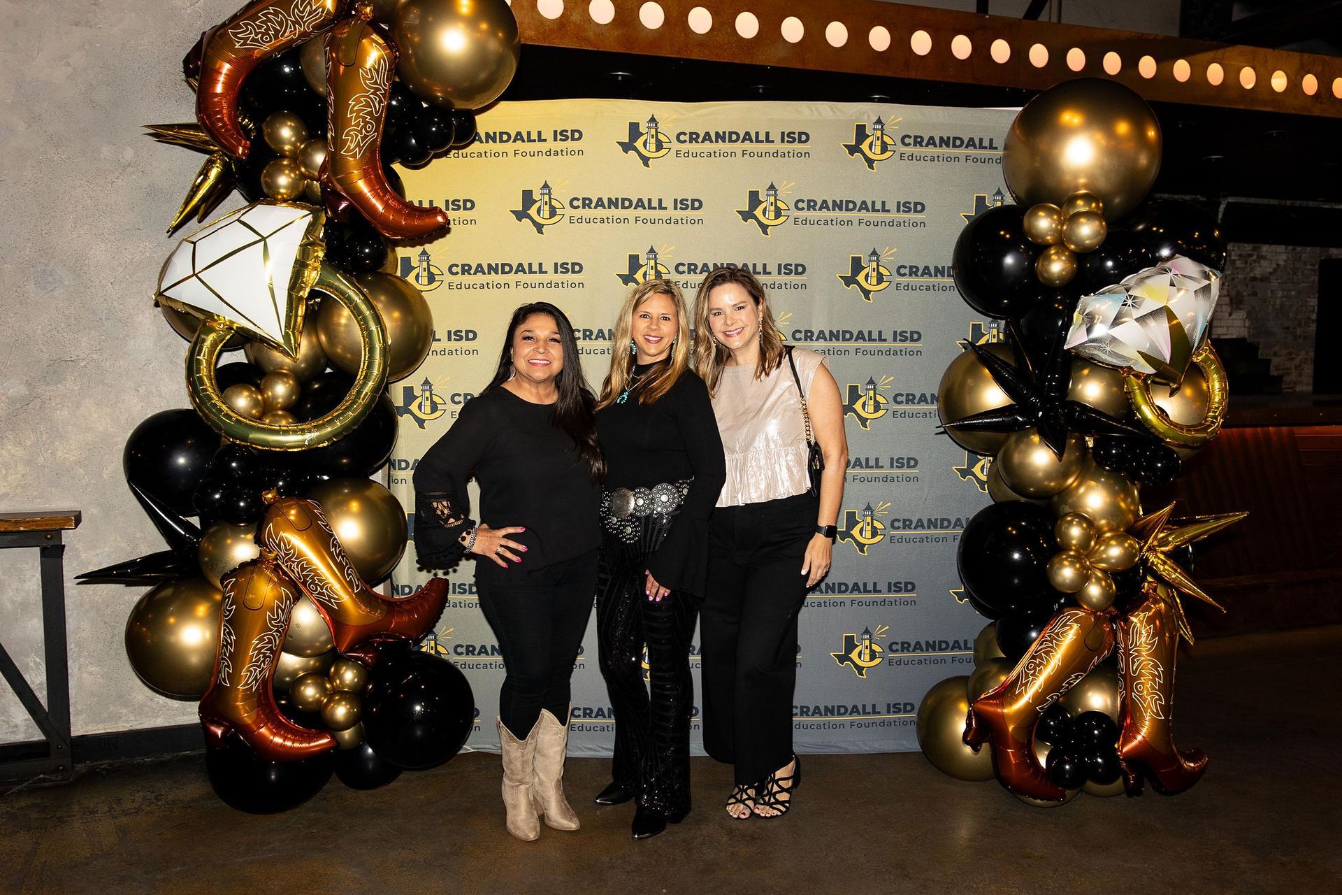Three women posing in front of a backdrop with gold and black balloons.