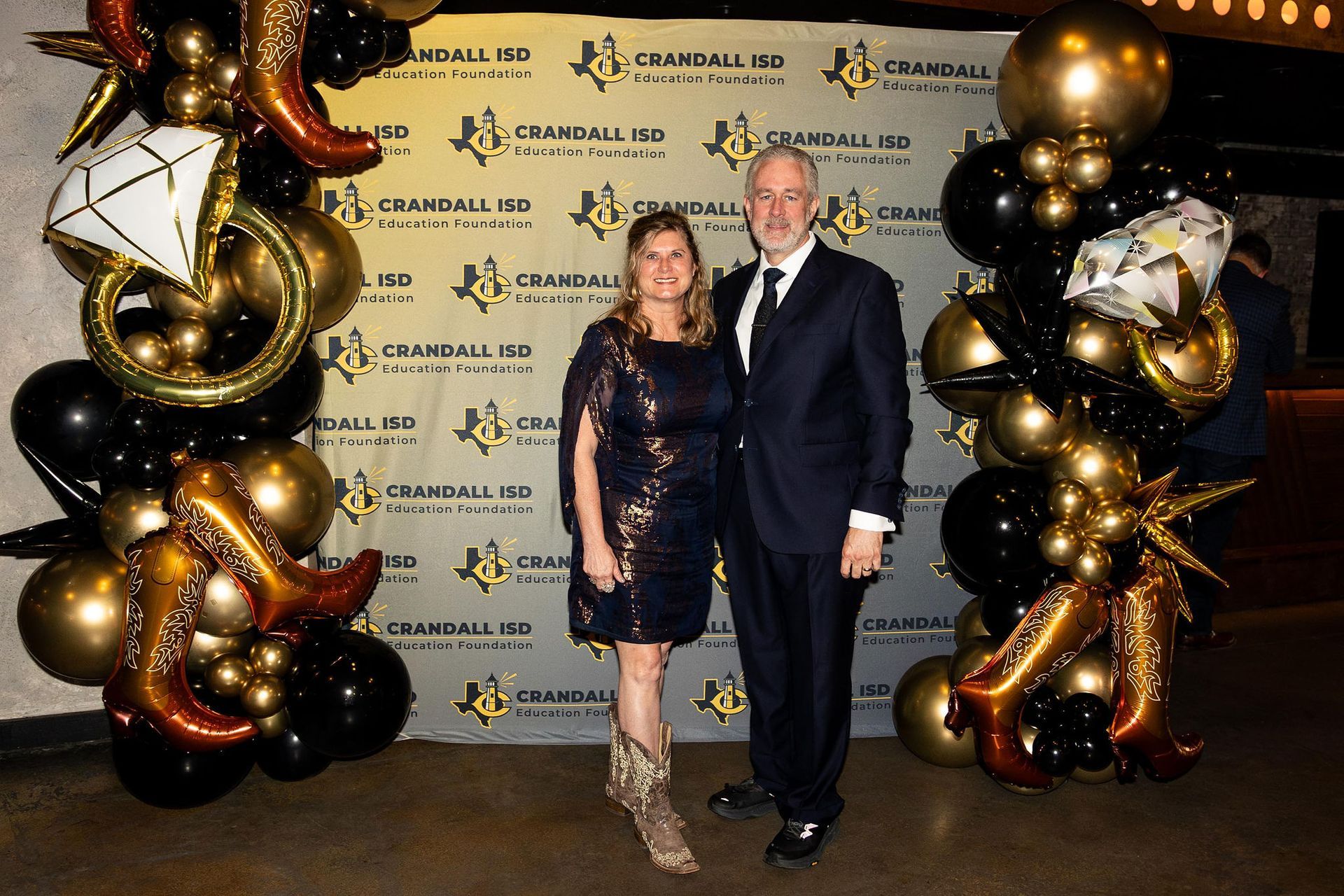 Couple posing in front of a backdrop at an event with balloons; woman in dress, cowboy boots, man in suit.