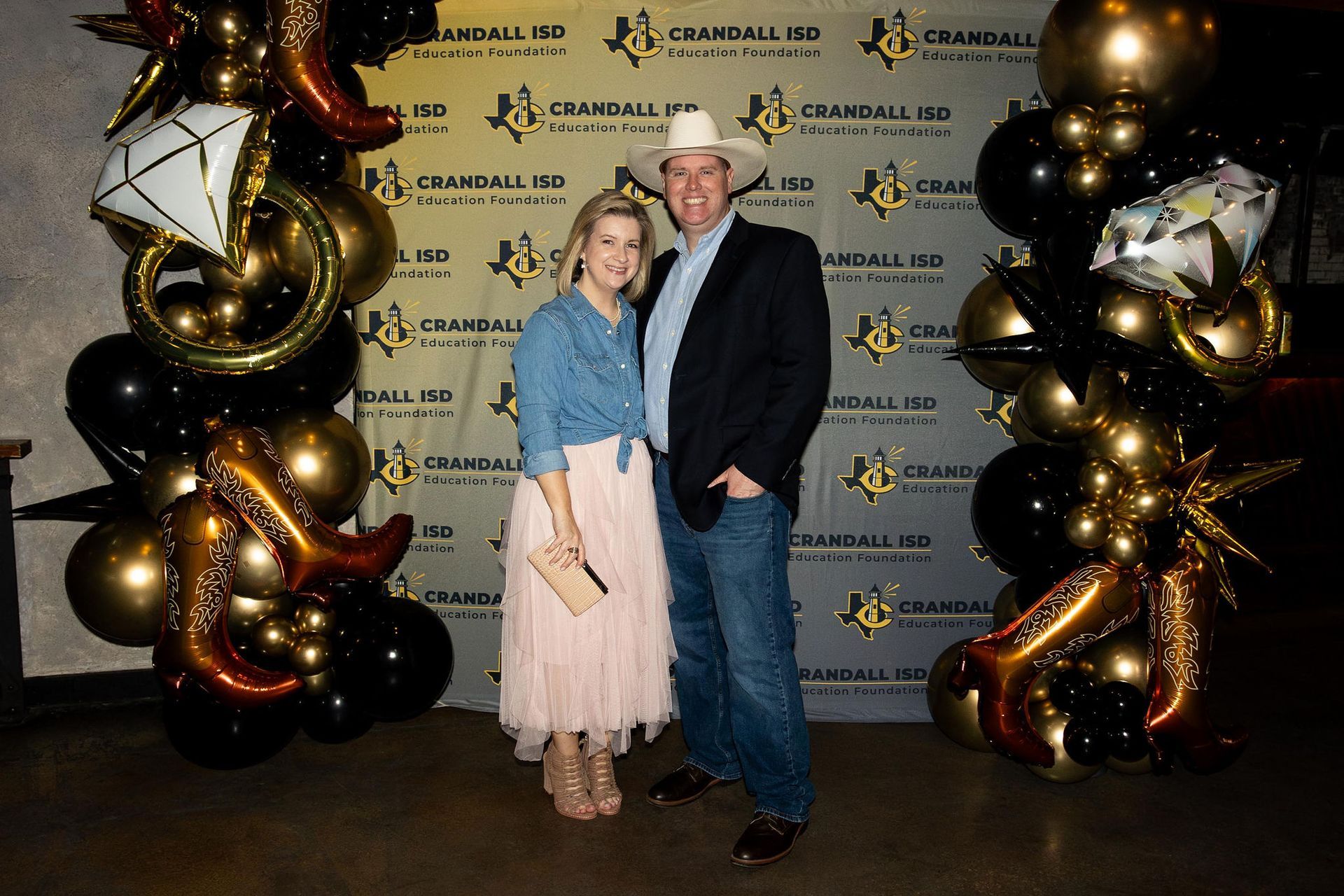 A couple poses at a formal event with a backdrop of balloons. The man wears a cowboy hat.
