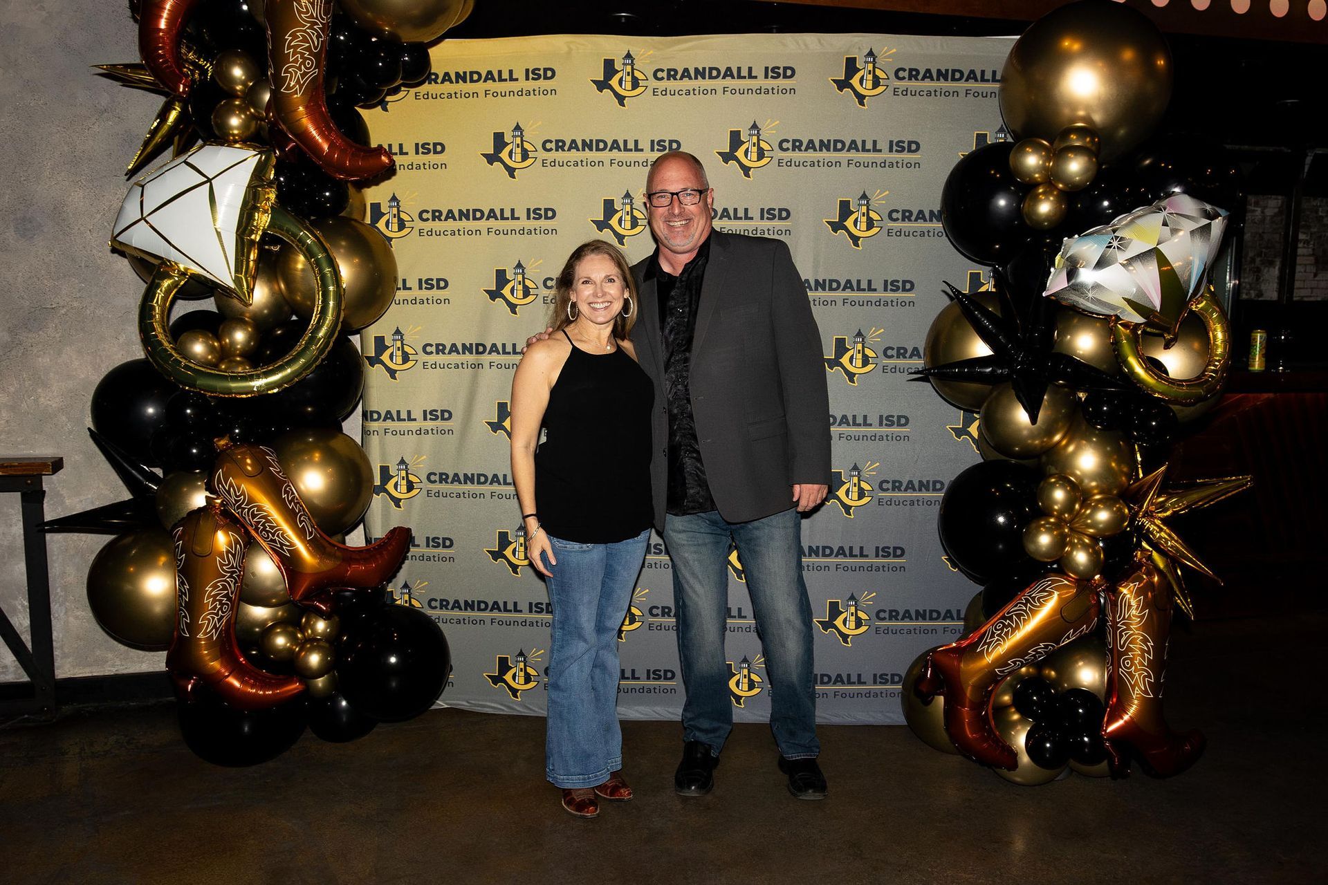 Woman and man posing in front of a backdrop with balloons. They are smiling and wearing casual clothes.