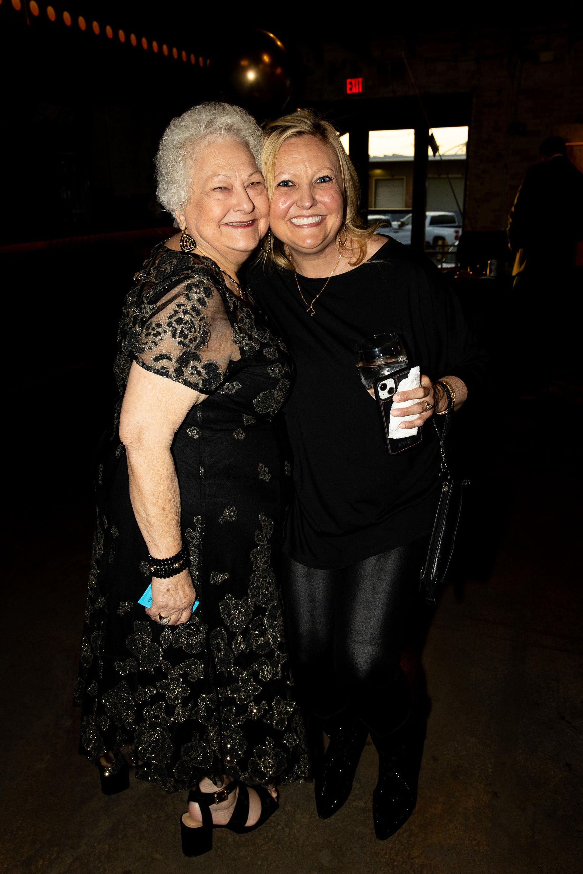 Two women embrace; one older in a black lace dress, the other younger in black pants and top, holding a drink.