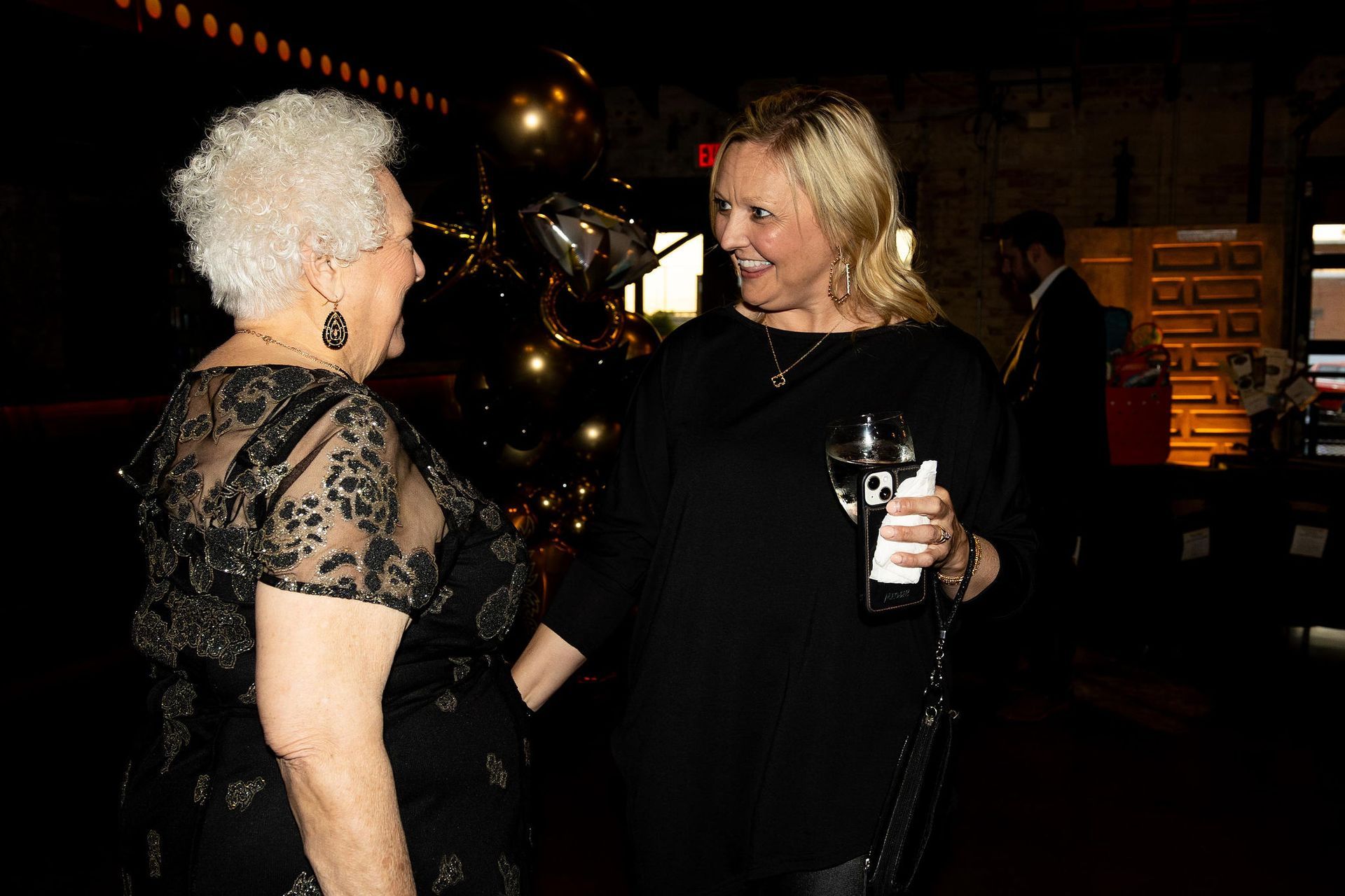 Two women in black dresses converse indoors; one holds a drink.