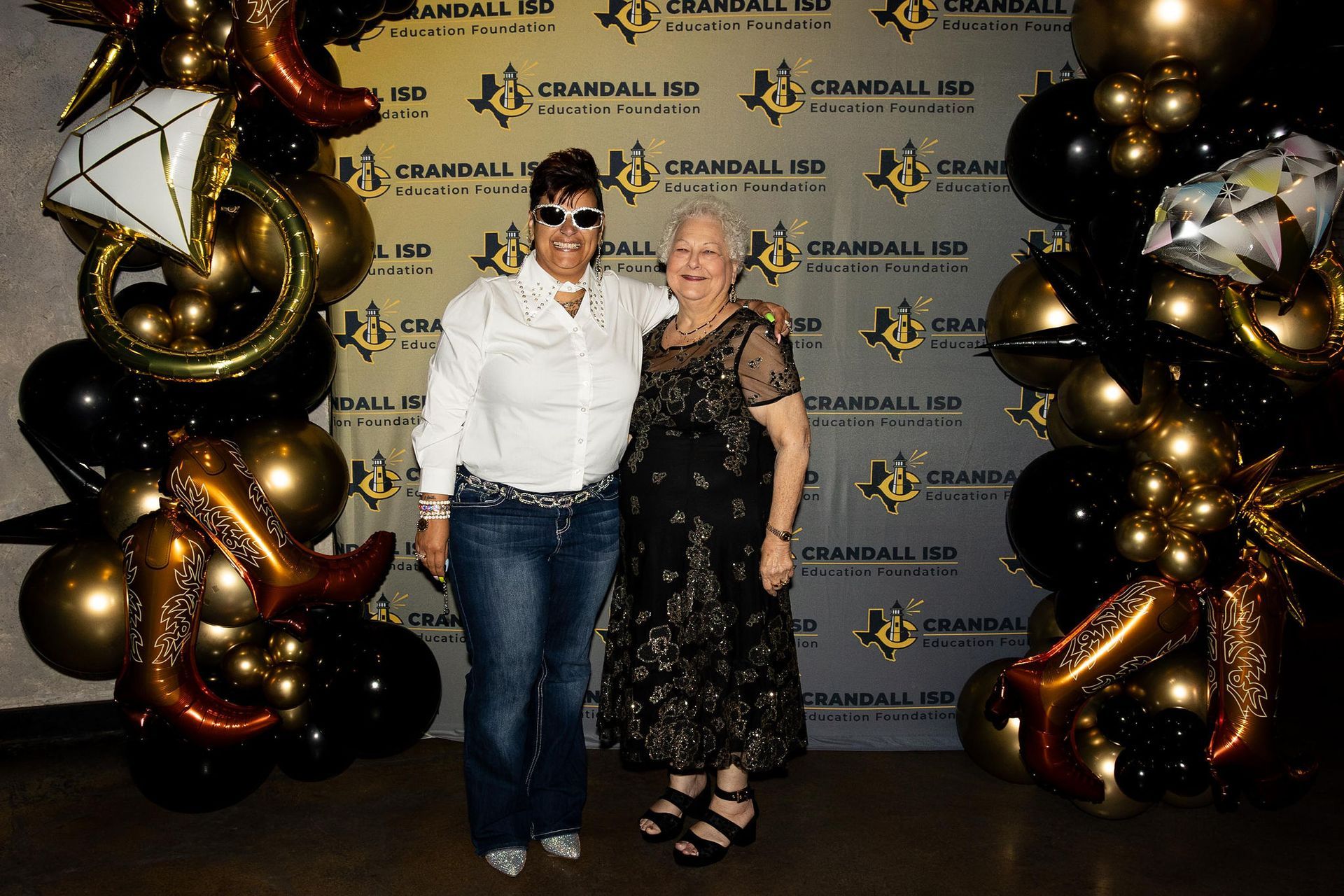 Two women pose, smiling, in front of a gold and black balloon arch and backdrop that reads 
