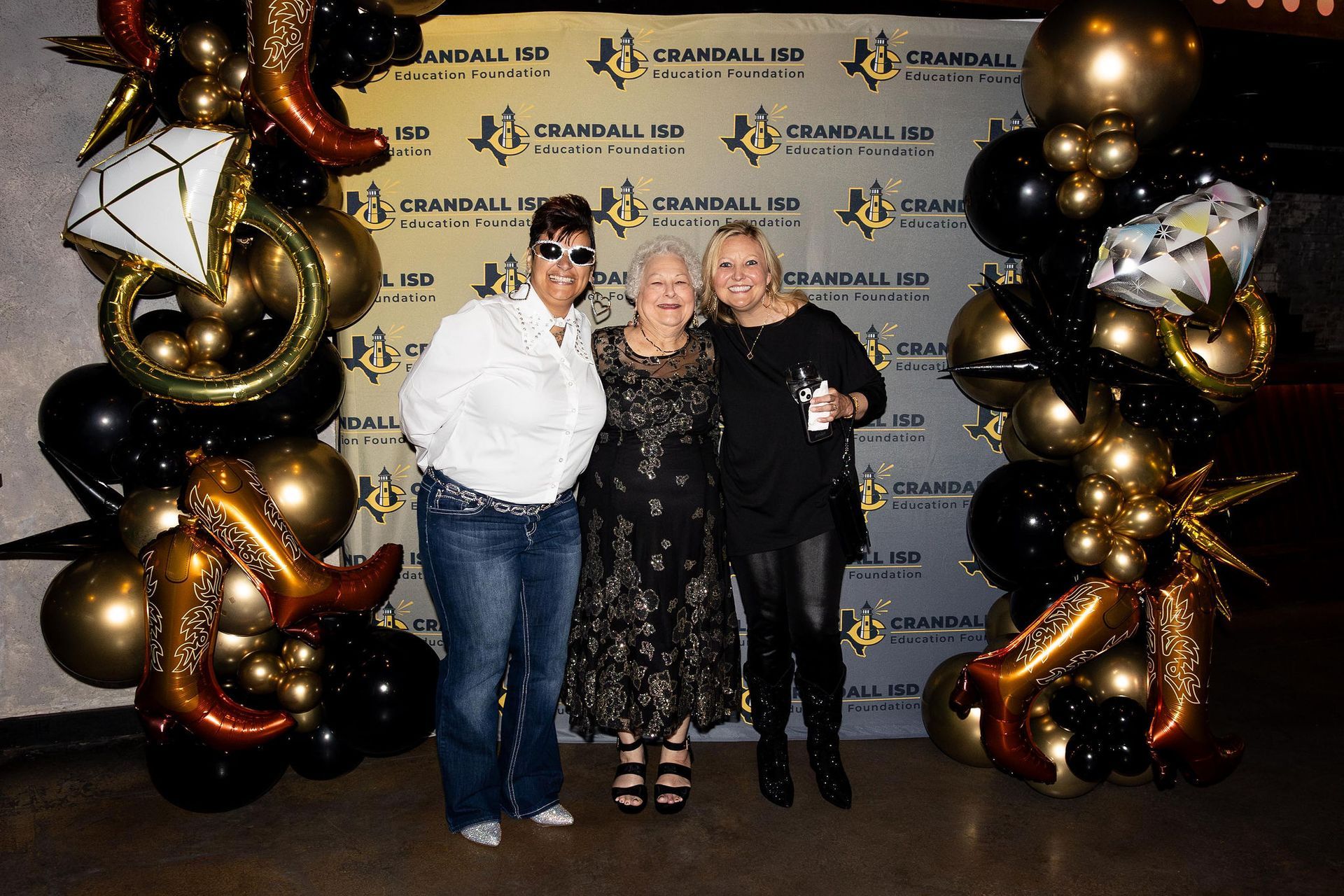 Three women at a party, posing in front of a backdrop with balloons.