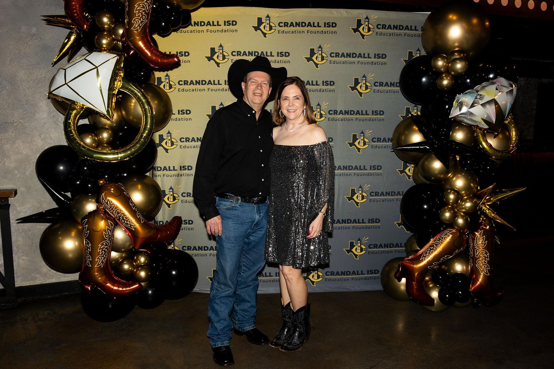A couple poses in front of a backdrop with black and gold balloons, she in a sequined dress, he in a cowboy hat.