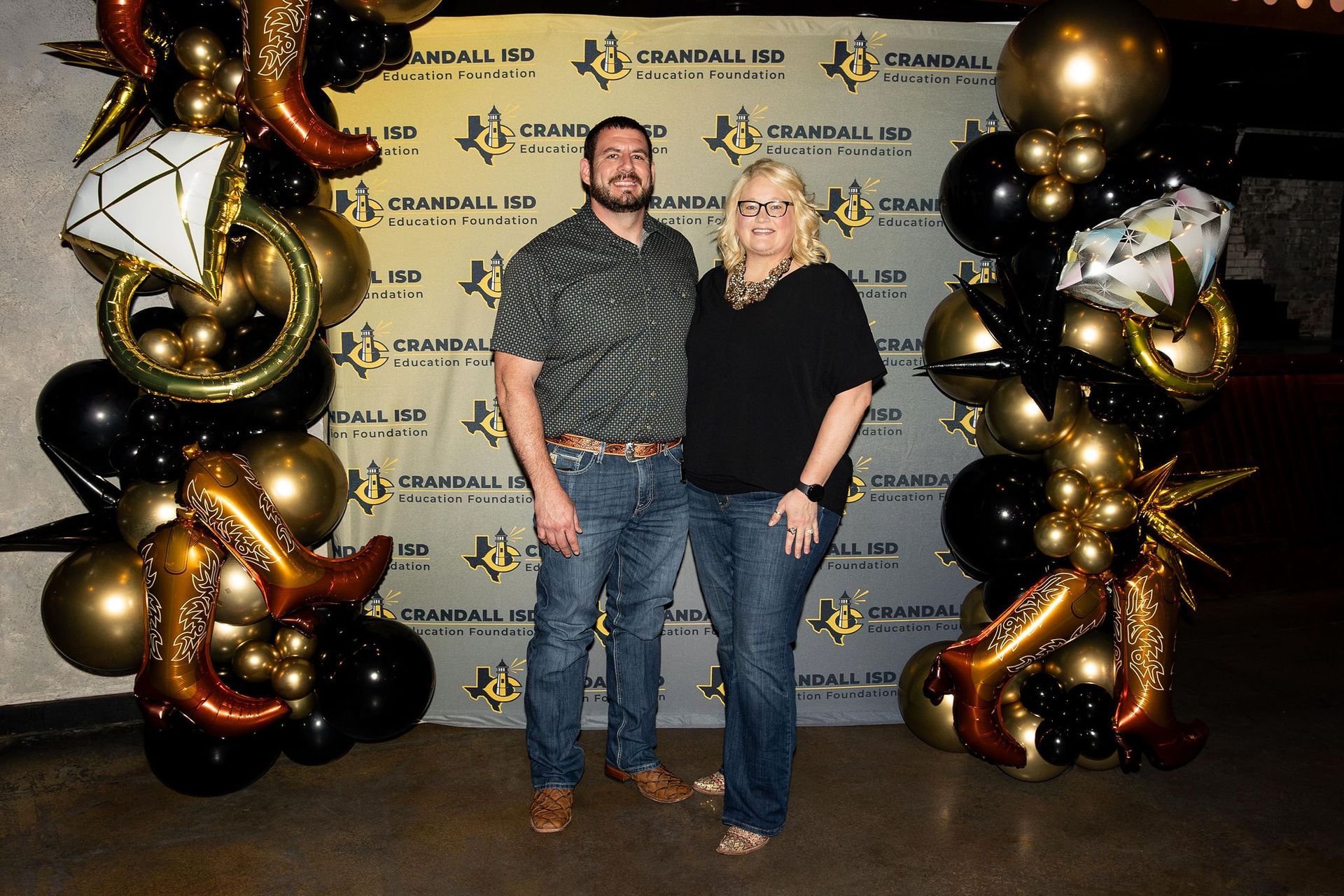 Couple posing in front of a step-and-repeat backdrop with balloons. Man in jeans and a patterned shirt. Woman in jeans and black top.