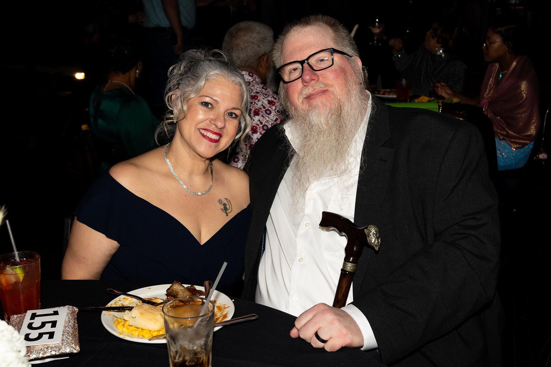 Couple at formal event, woman in blue dress smiling, man with beard and cane, seated at table.