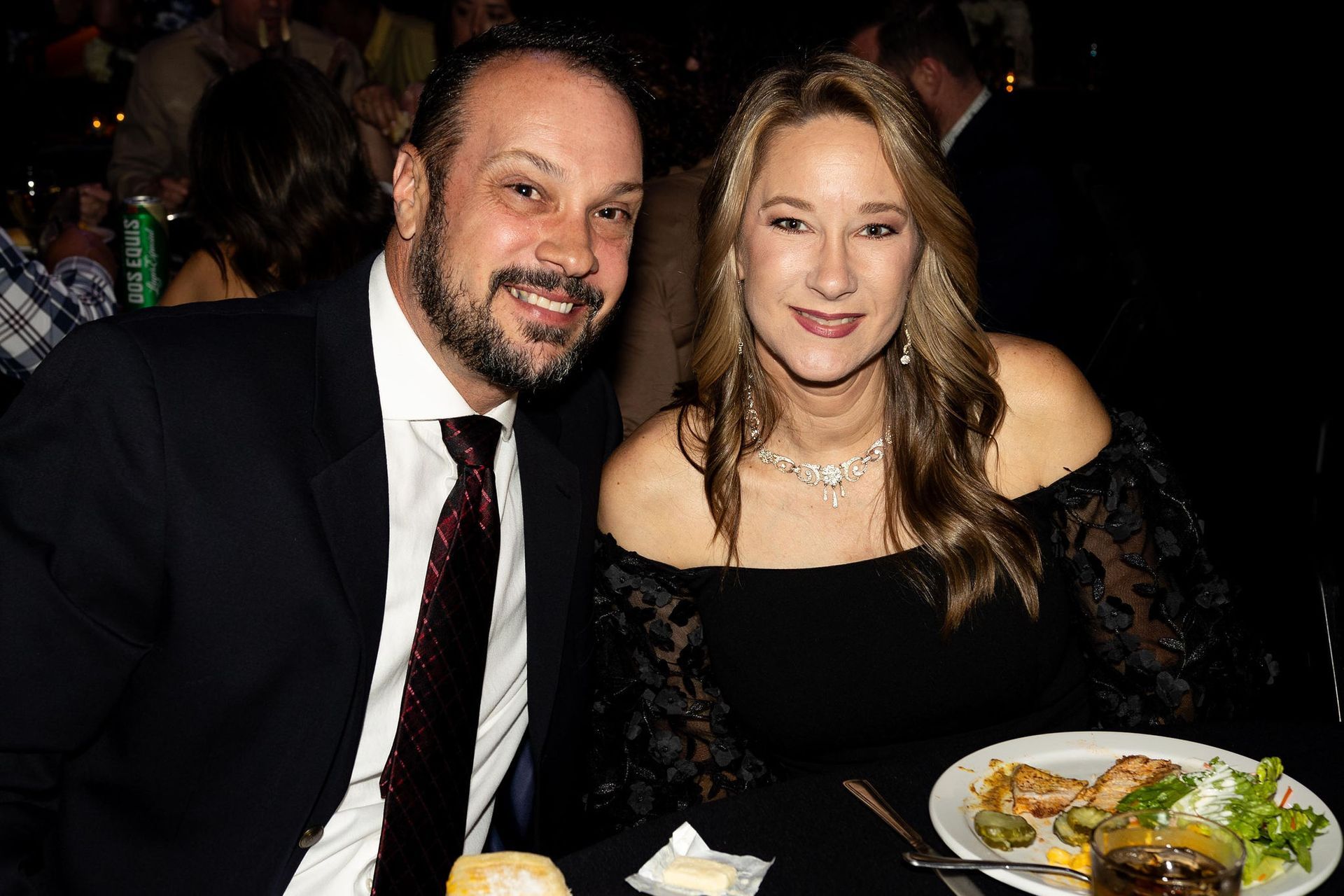 Man and woman seated at a table, smiling at the camera. Man in suit, woman in black dress with necklace.