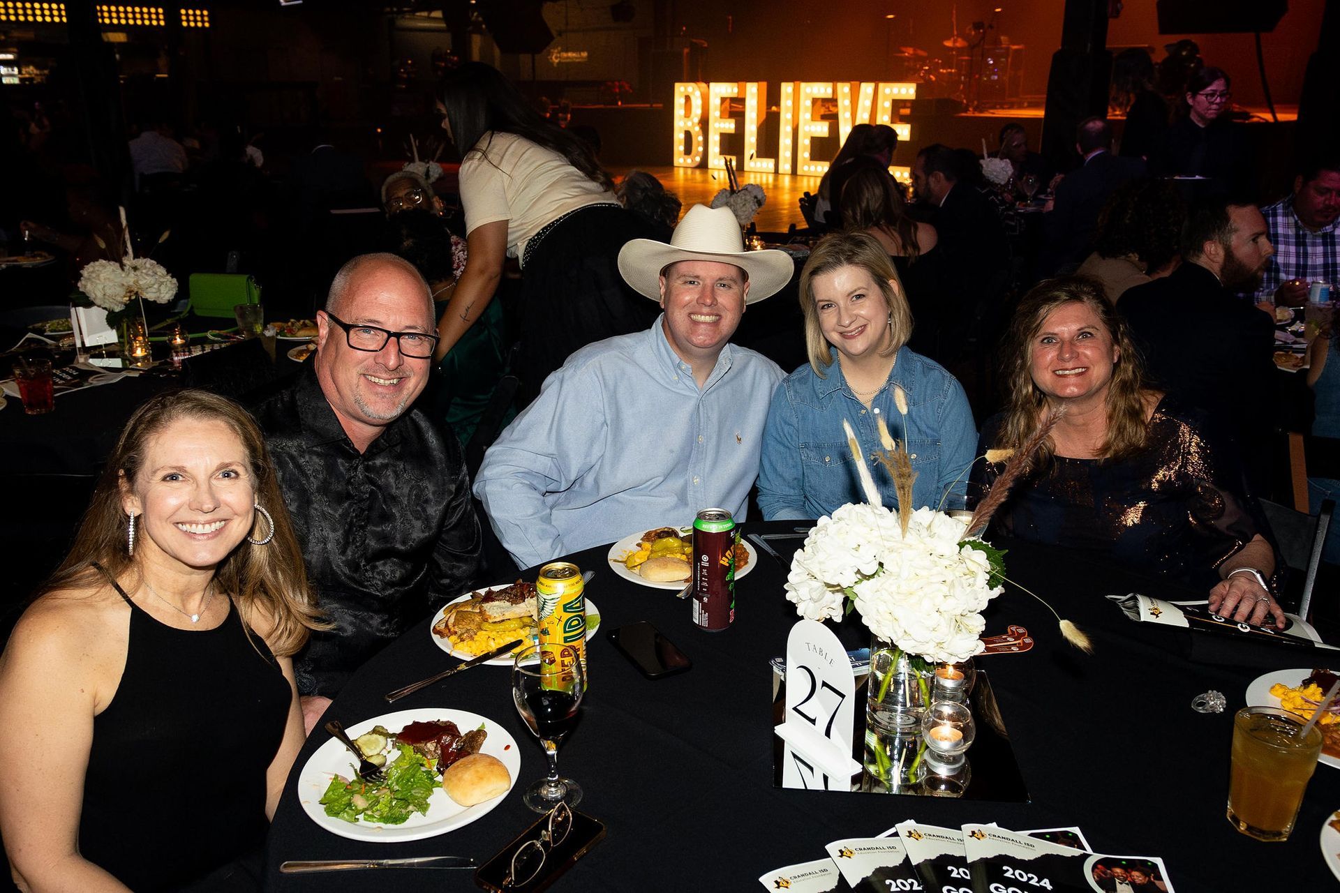 Group of people at a formal dinner seated at a table with food, drinks, and a 