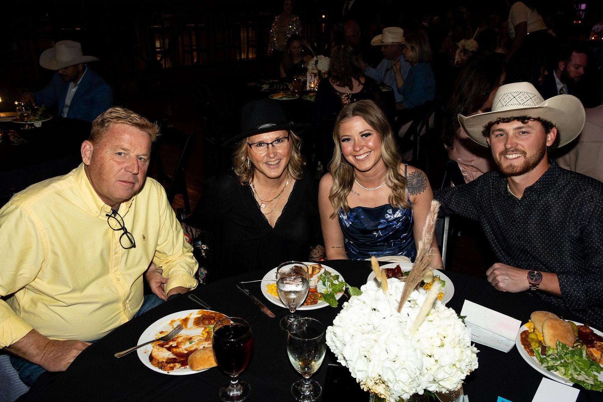 Four people at a dinner table at a formal event, smiling, eating. Two are wearing cowboy hats.