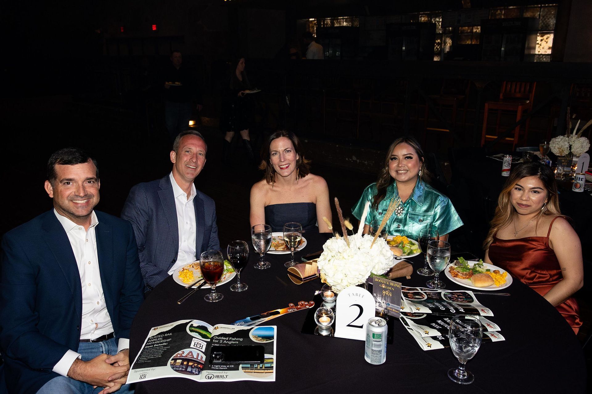 Five people seated at a table, dining.  Dark setting.  Woman in turquoise, and another in rust dress.