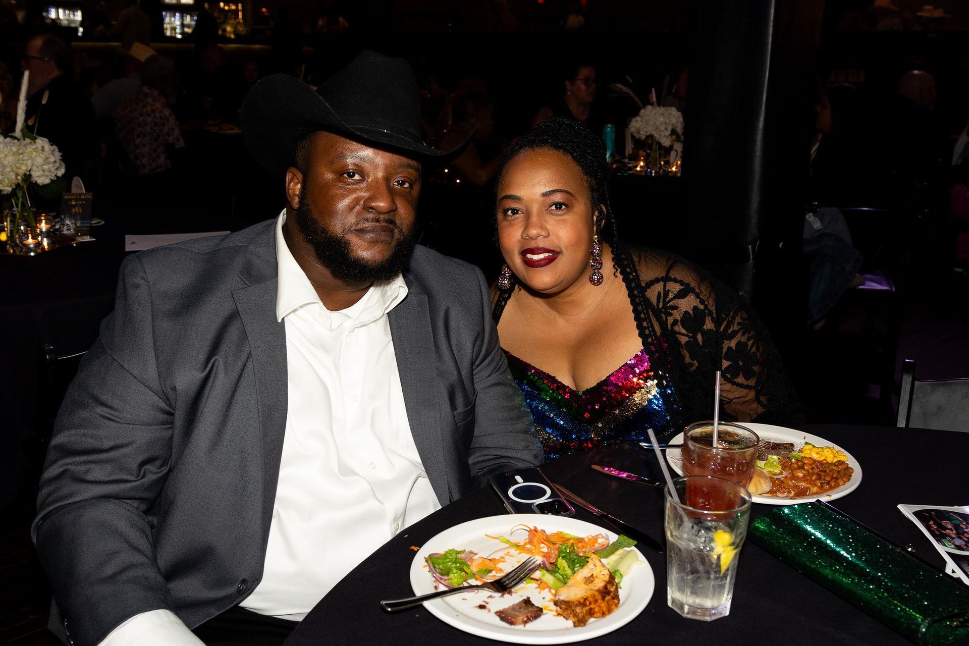 A Black couple smiles at a table with food and drinks. Man in a suit and hat, woman in sequin top.