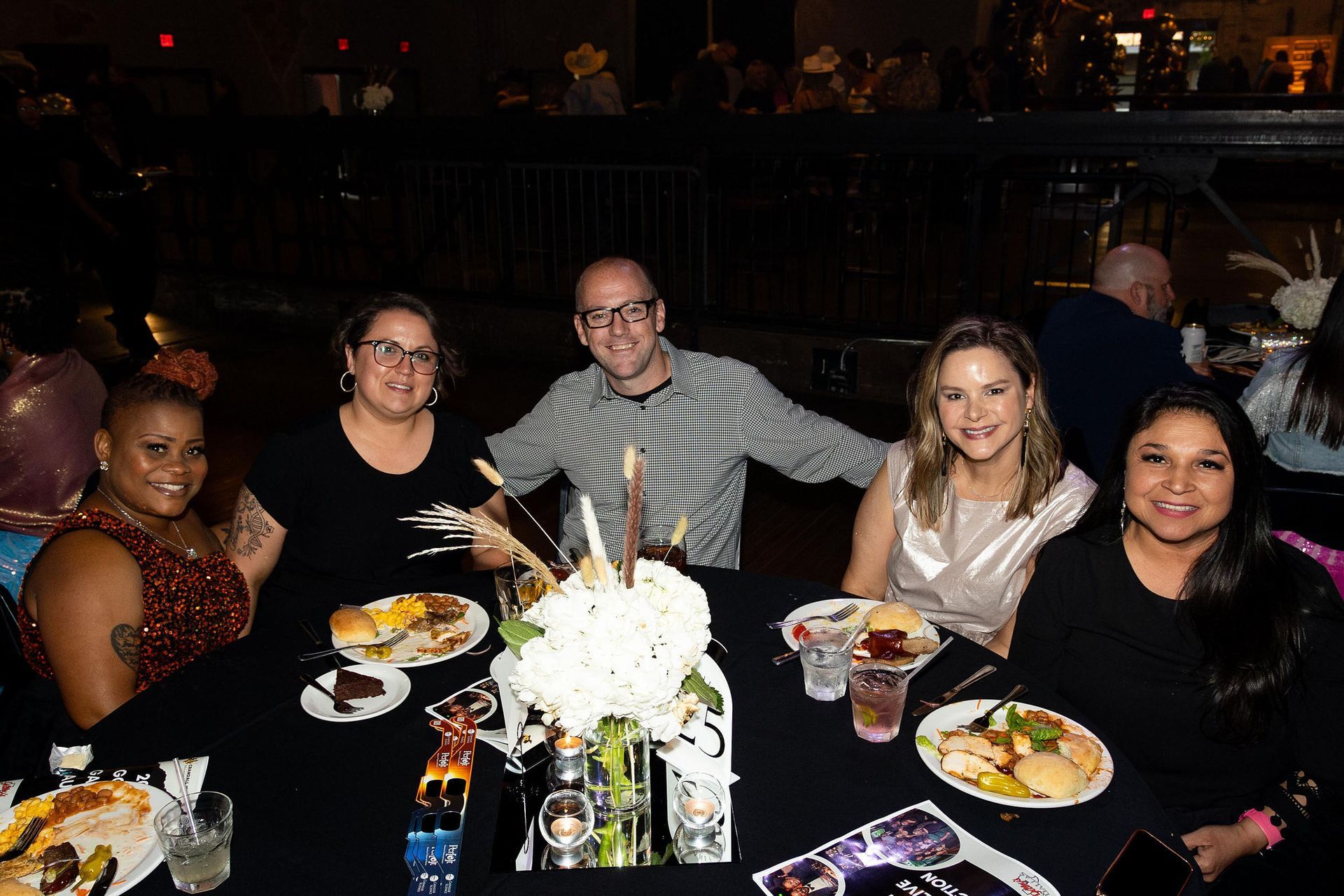Group of five people smiling at a table set for dinner, with food and a flower centerpiece.