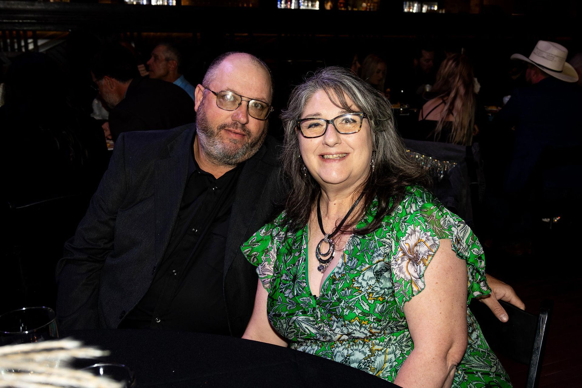 A smiling couple sits together, the man in a dark suit and the woman in a green floral dress.