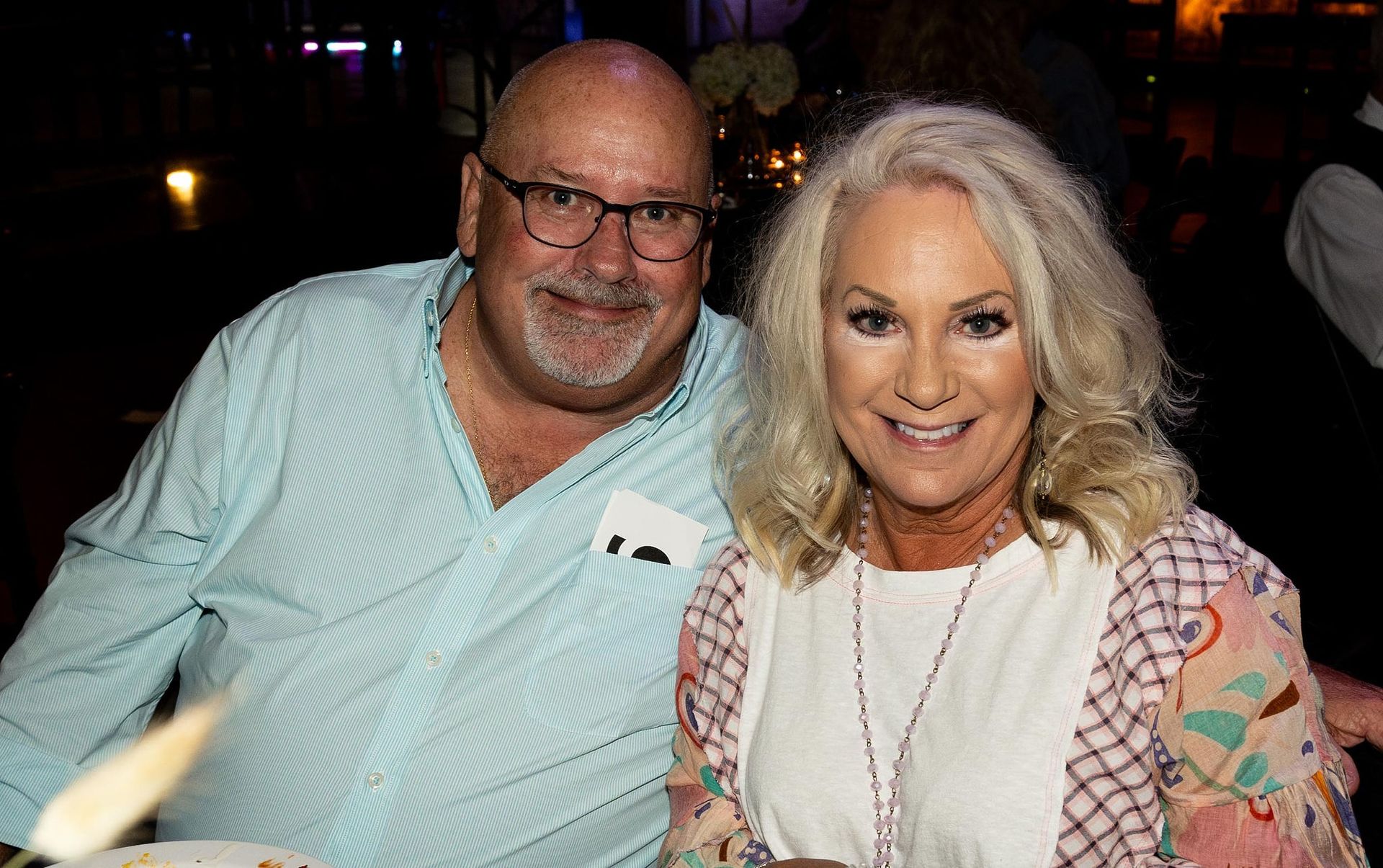 Couple smiling at camera; man in glasses, light blue shirt; woman with blonde hair, floral sleeves.