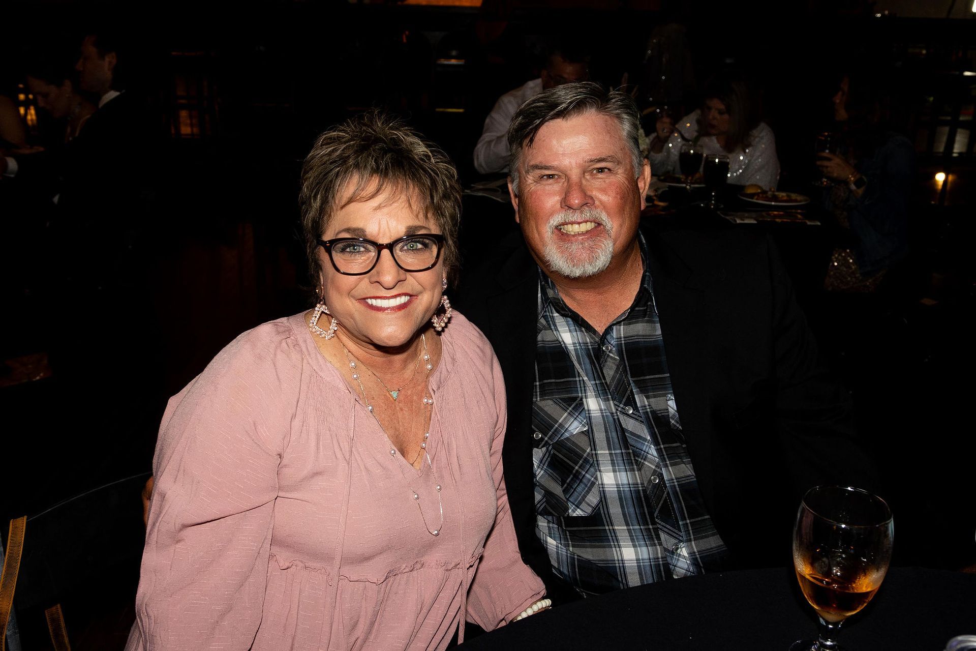 Smiling couple at a dimly lit restaurant. Woman in pink top, man in plaid shirt and blazer.