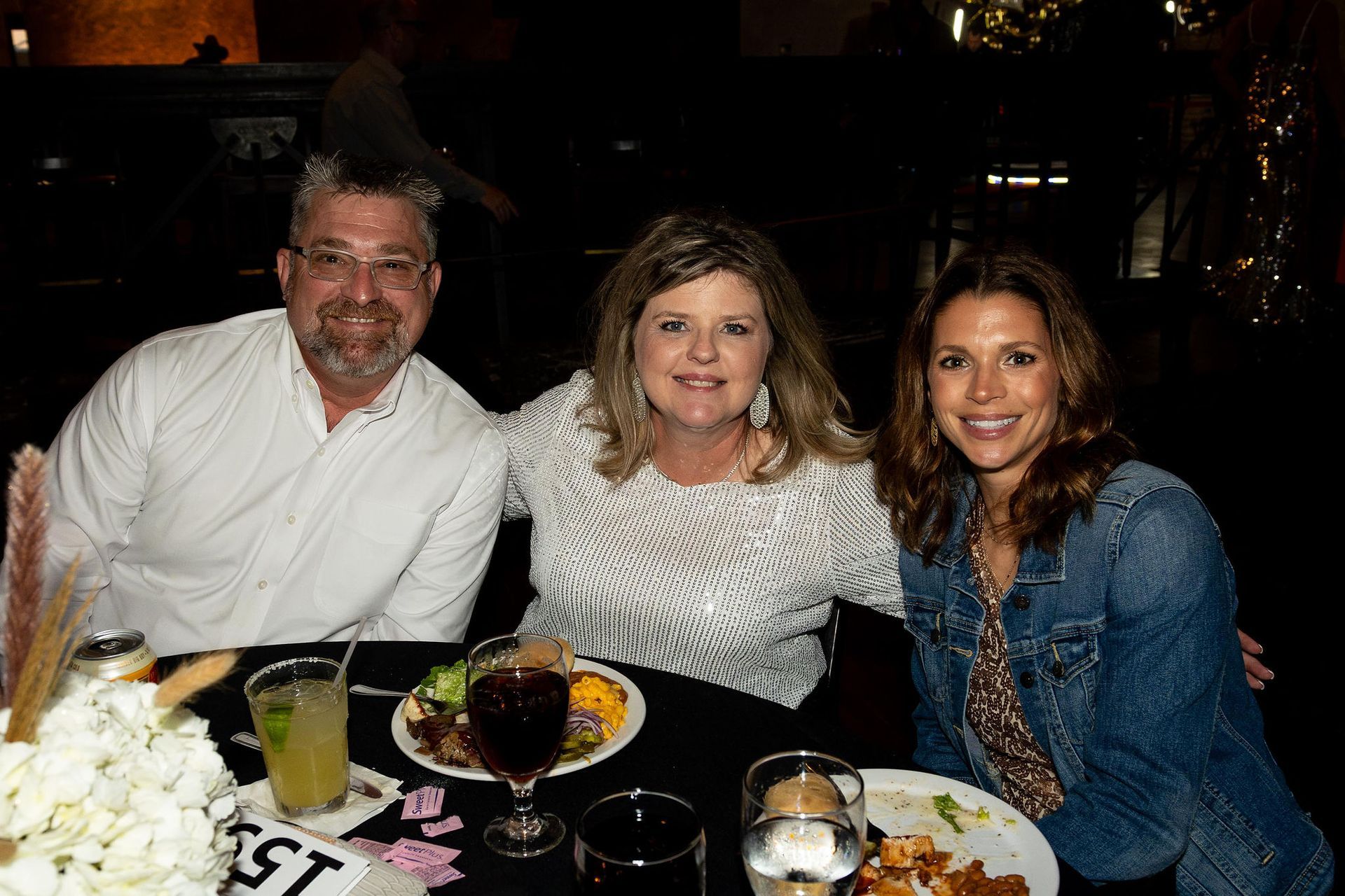 Three smiling people at a table. Man with glasses, woman in white top, woman in denim jacket. Restaurant setting.