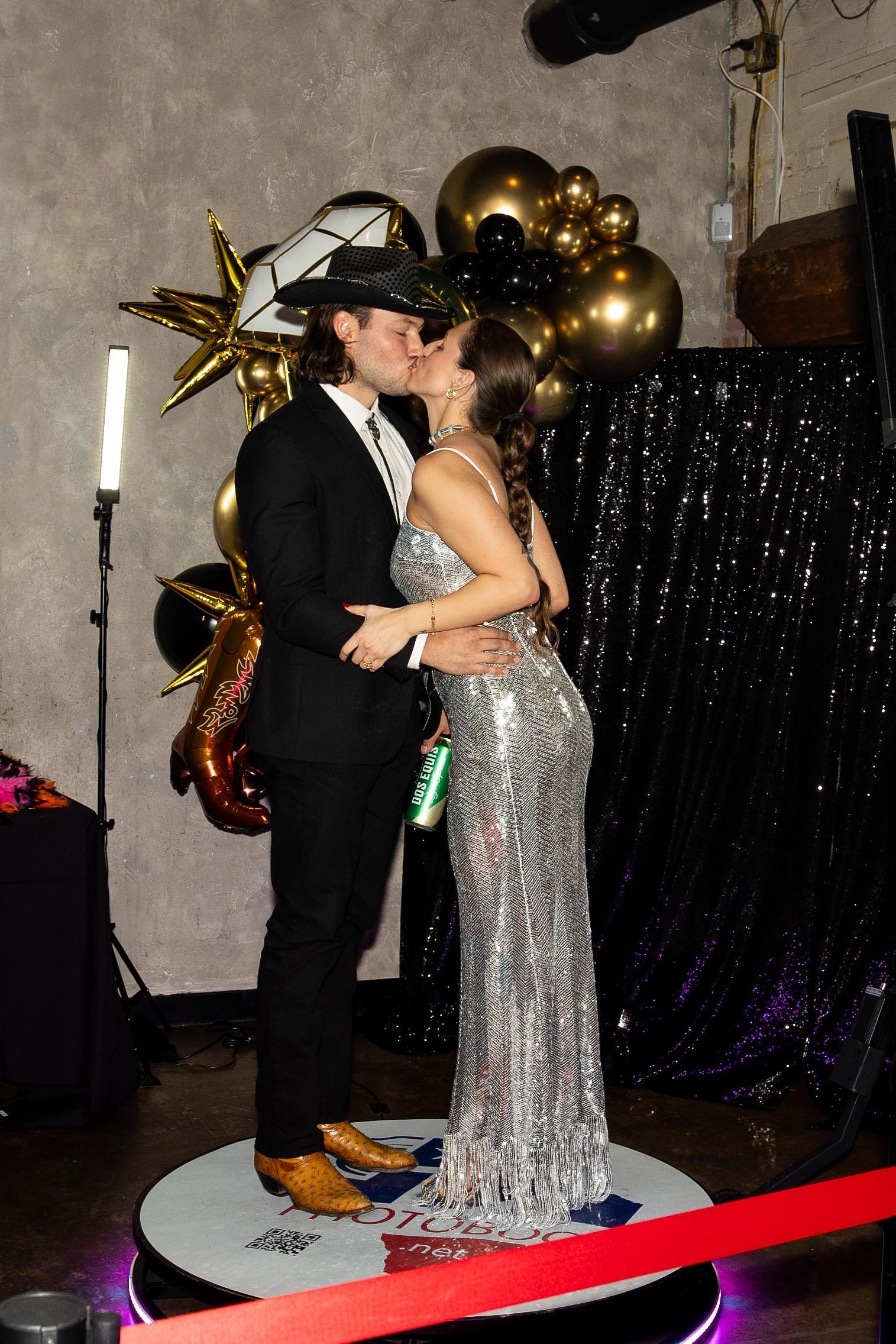 A couple kissing on a rotating photo booth platform, dressed in formal attire, at a party.