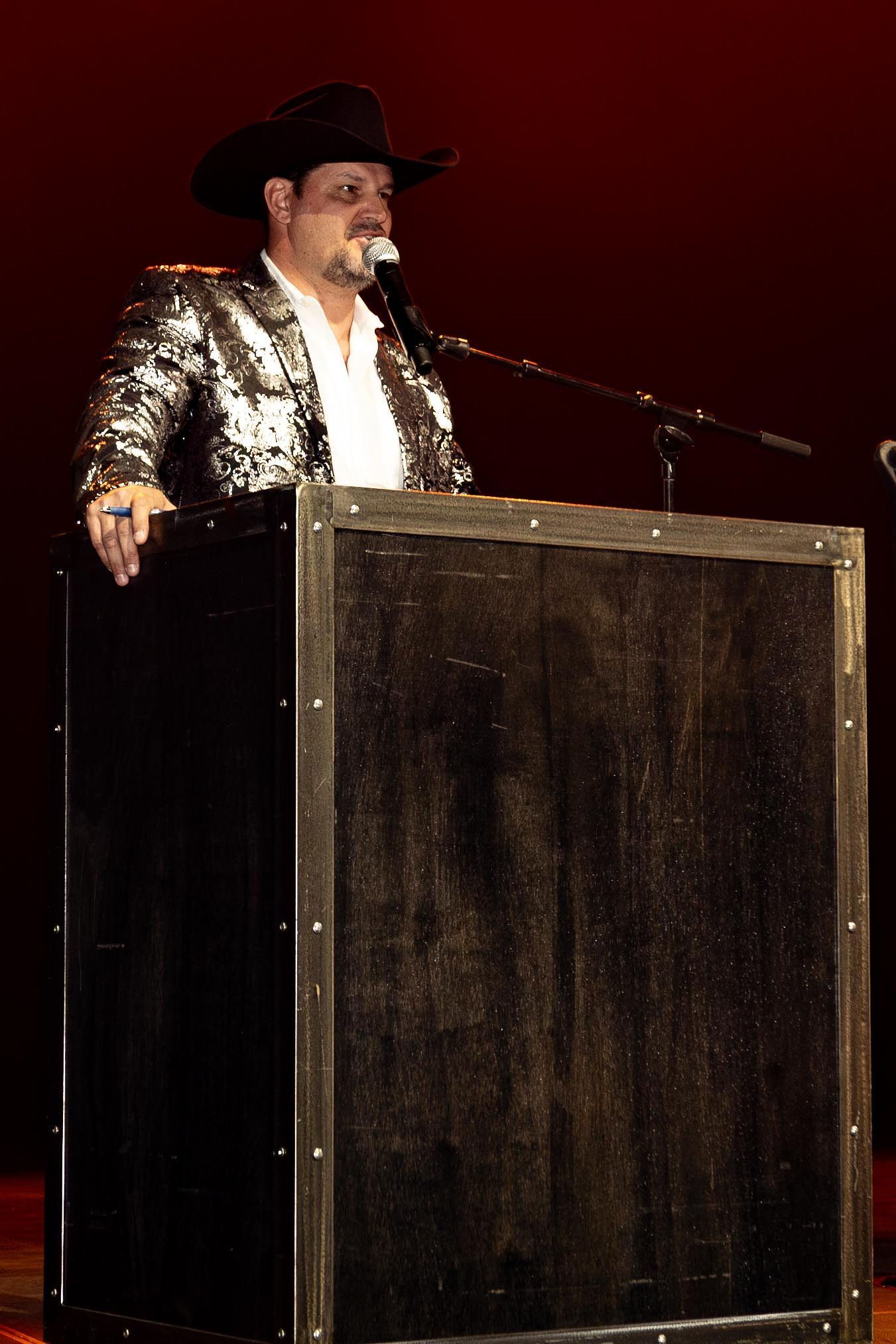 Man in sequined jacket, cowboy hat, speaking at a podium on a stage, microphone in front.