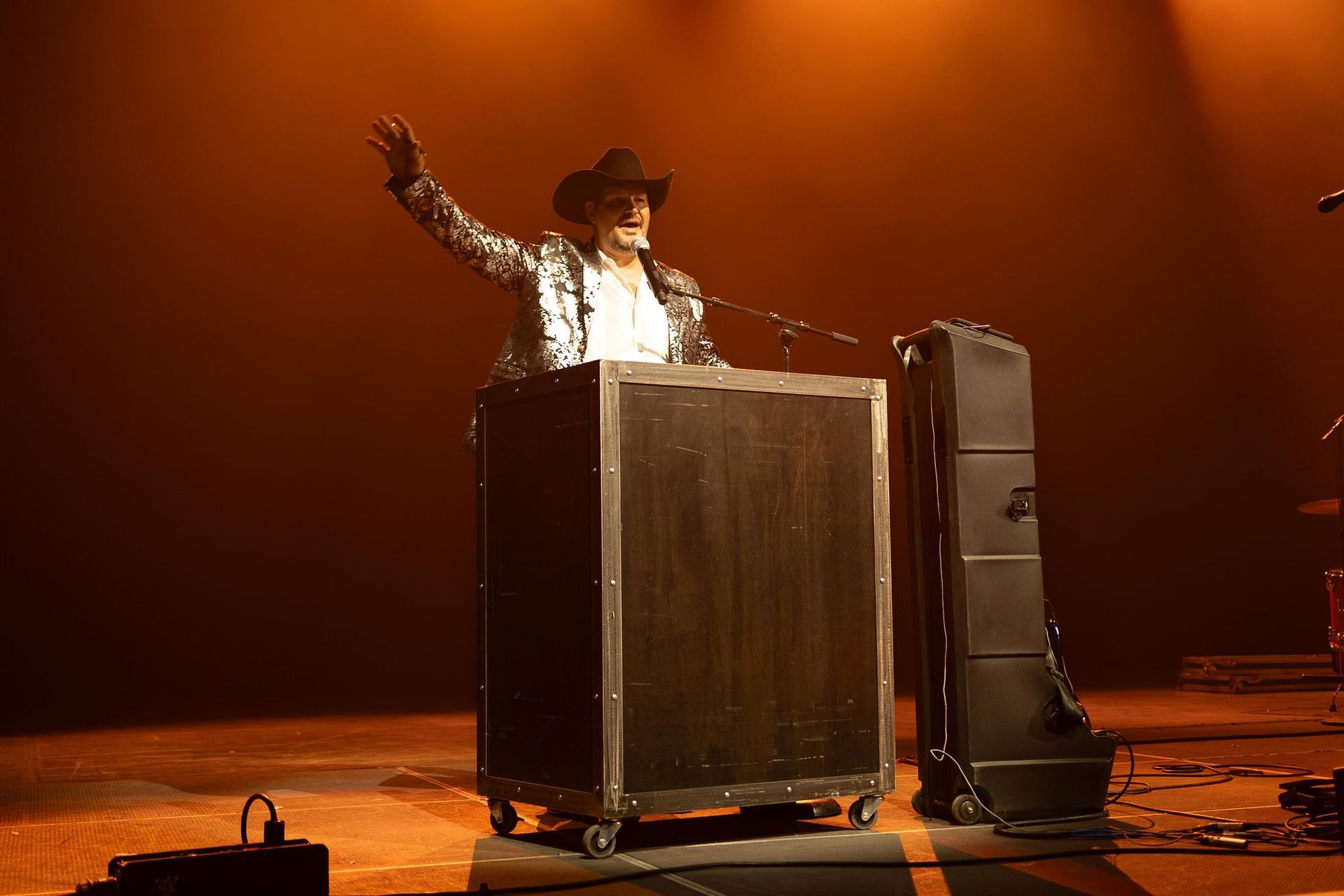 Person in sequined jacket and cowboy hat on stage, gesturing; behind a large speaker box; stage bathed in orange light.