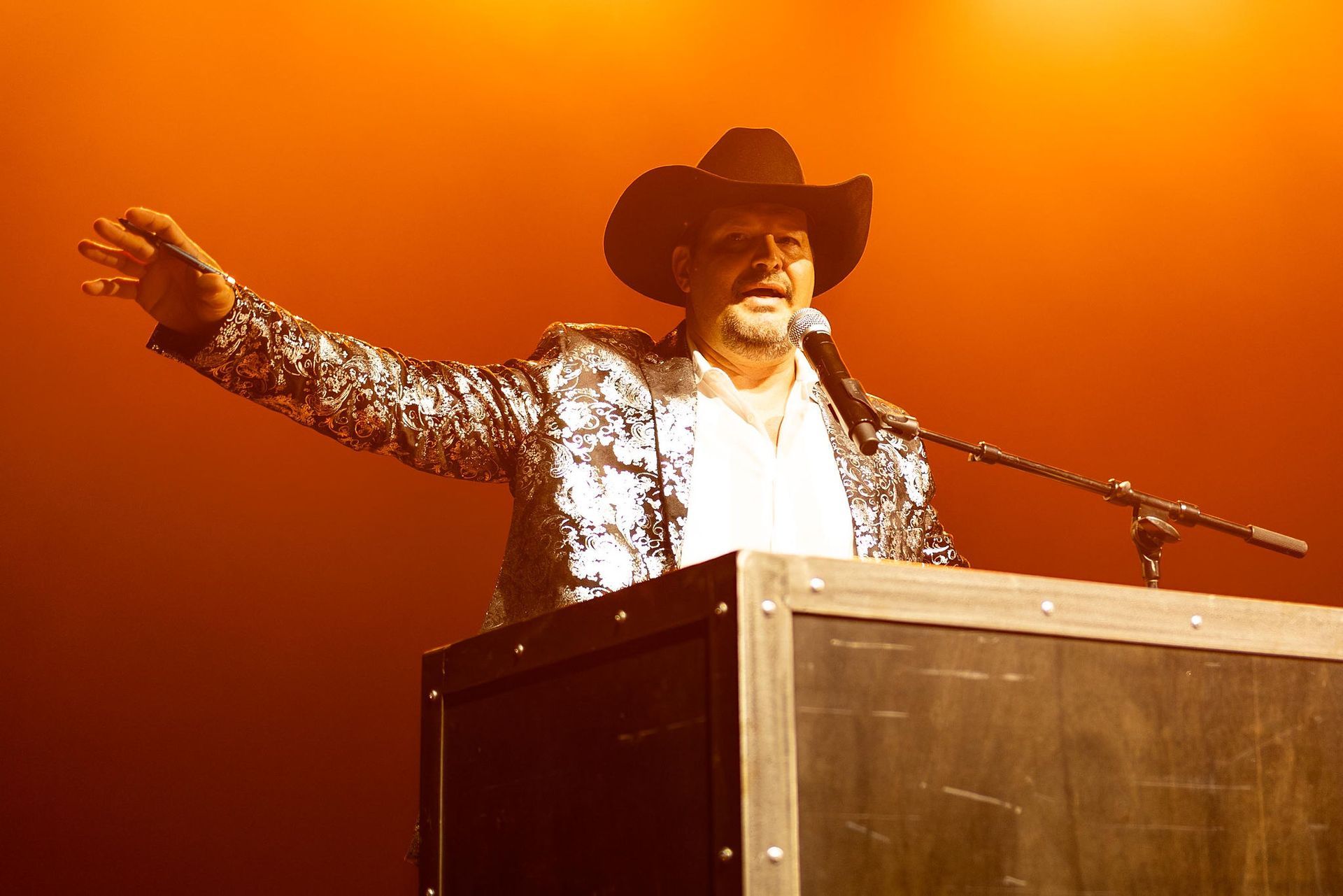 Man in a cowboy hat, sparkly jacket, at a podium, speaking with a raised hand, warm lighting.